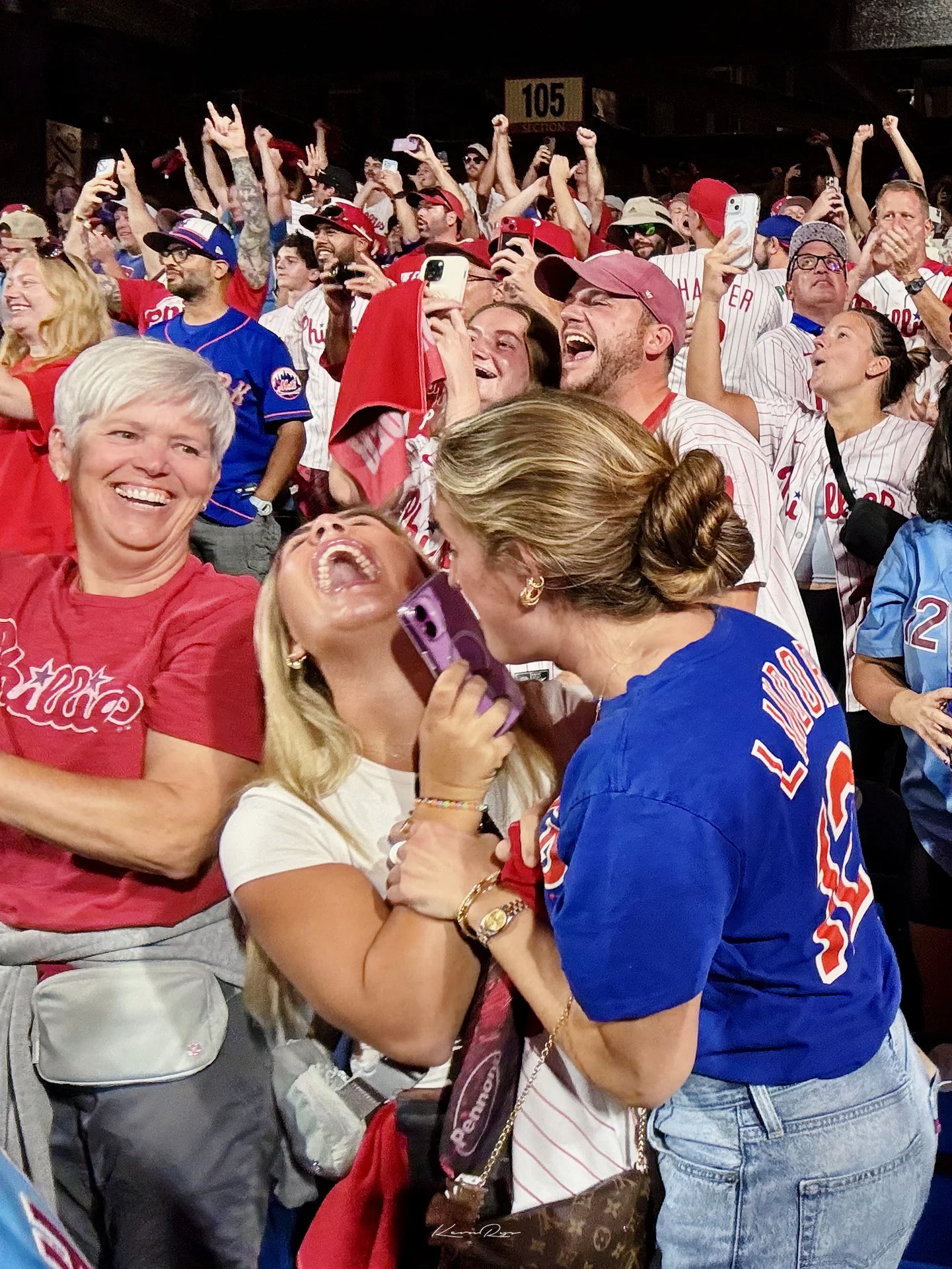 Fans in red and pinstripes scream with joy in the stands, phones raised, two in front laughing with their heads back