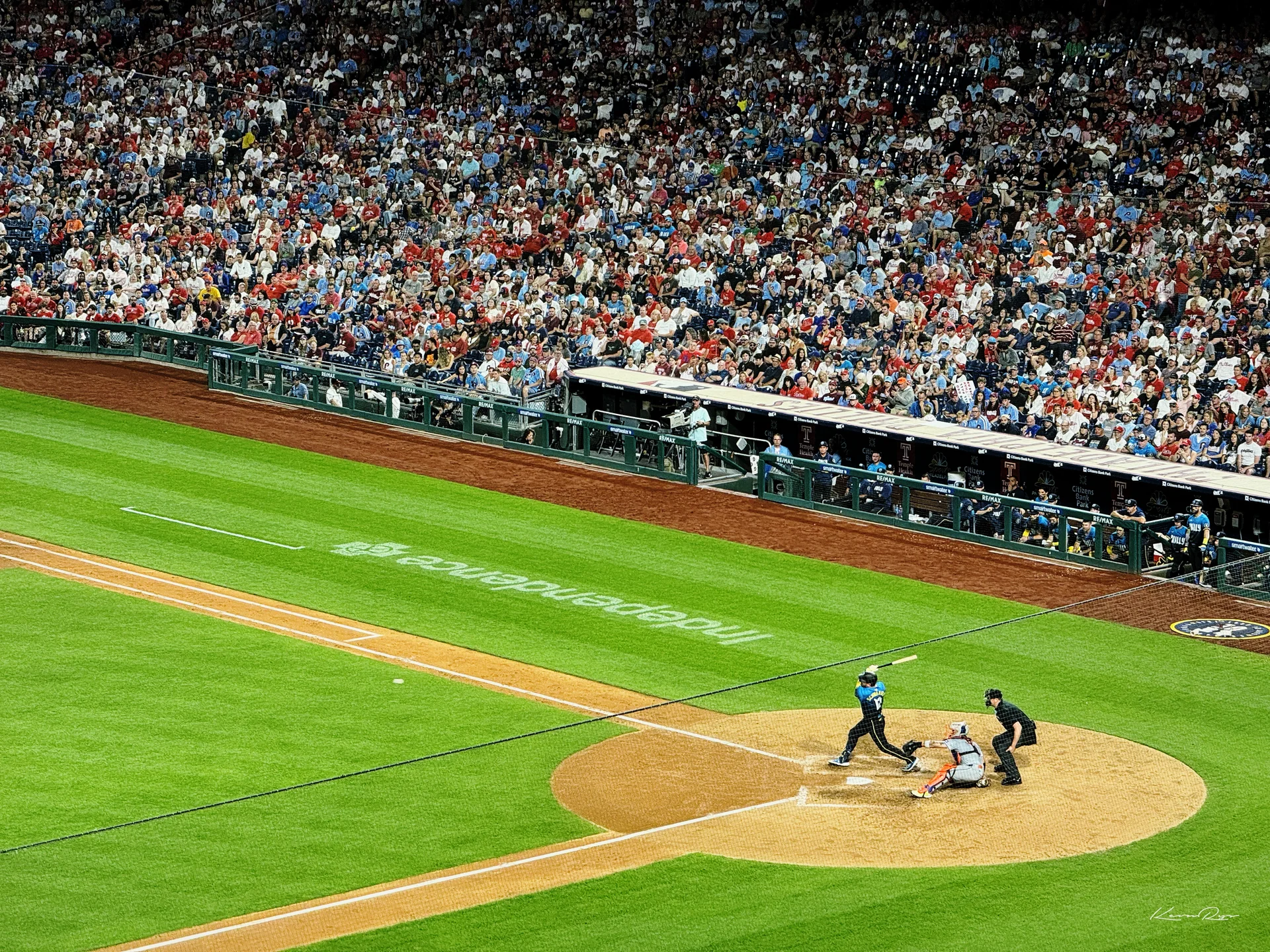 A batter in a light blue uniform swings at home plate at night as the catcher crouches, the stands packed behind the dugout