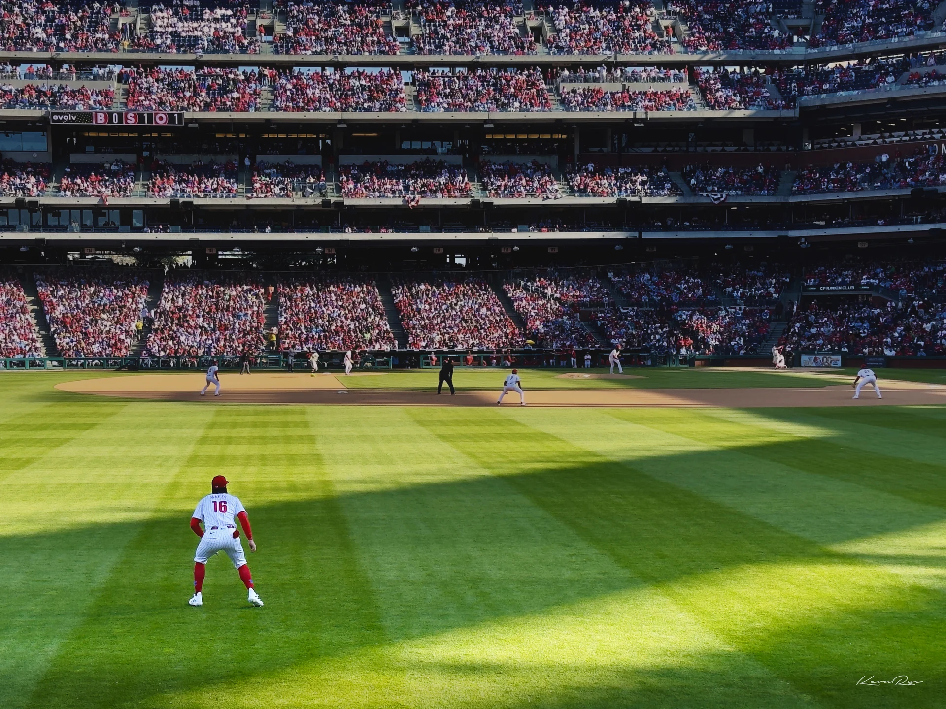 A Phillies outfielder in white with red socks waits in the grass as stadium shadows stretch across the field toward a full crowd