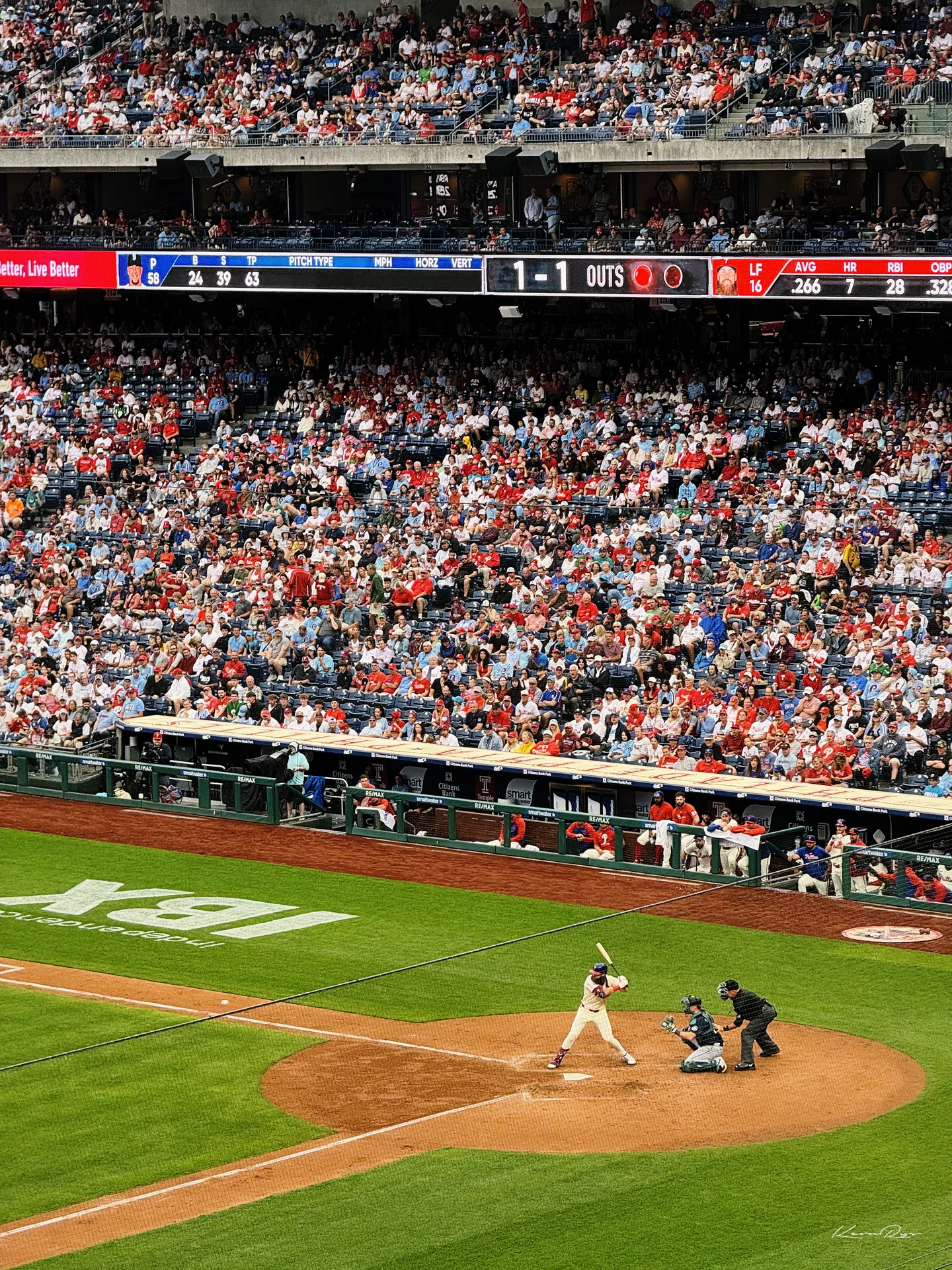 A batter waits for a pitch at home plate under the scoreboard, the catcher crouched behind with the stands packed in red