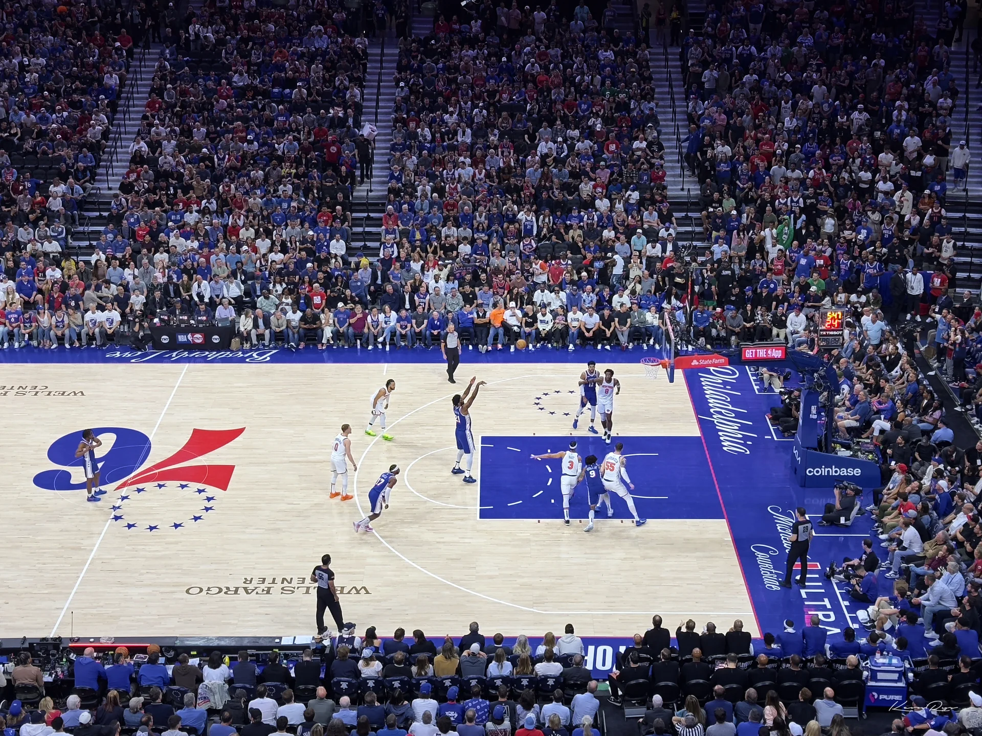 A player rises for a jump shot from beyond the arc as teammates crowd the paint, a packed arena watching from above