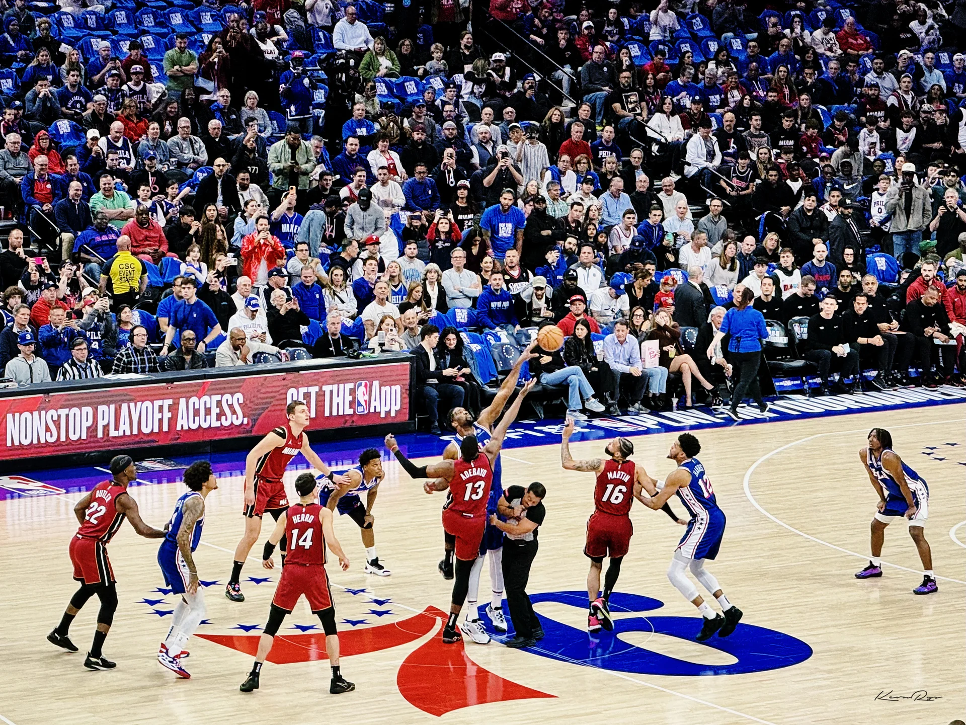 A Heat player and a Sixers player leap for the ball at center court, a packed crowd in blue filling the stands behind them