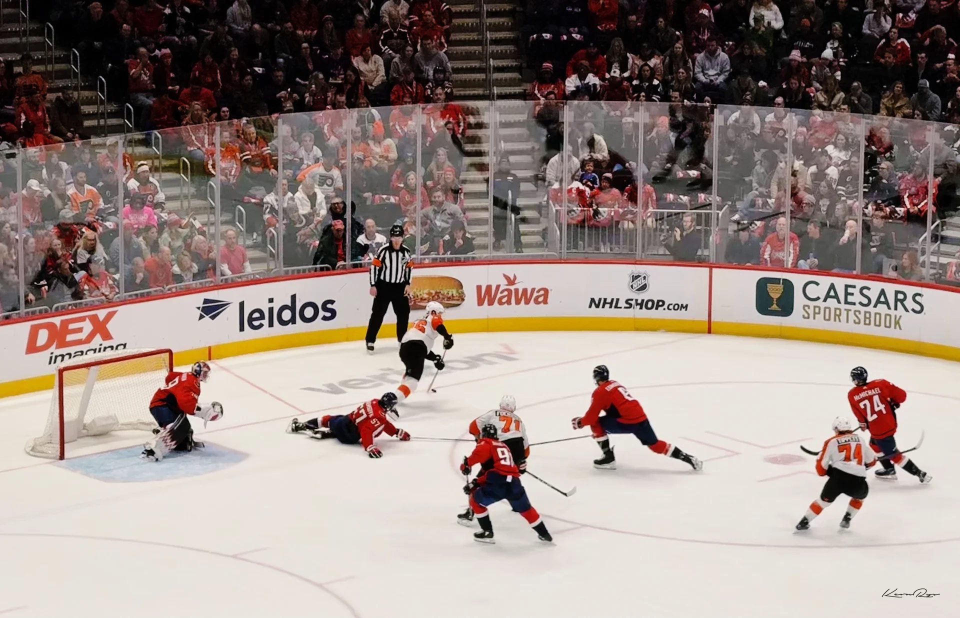 A Flyers skater in white cuts toward the net as a Capitals player slides on the ice, the goalie crouched in the crease