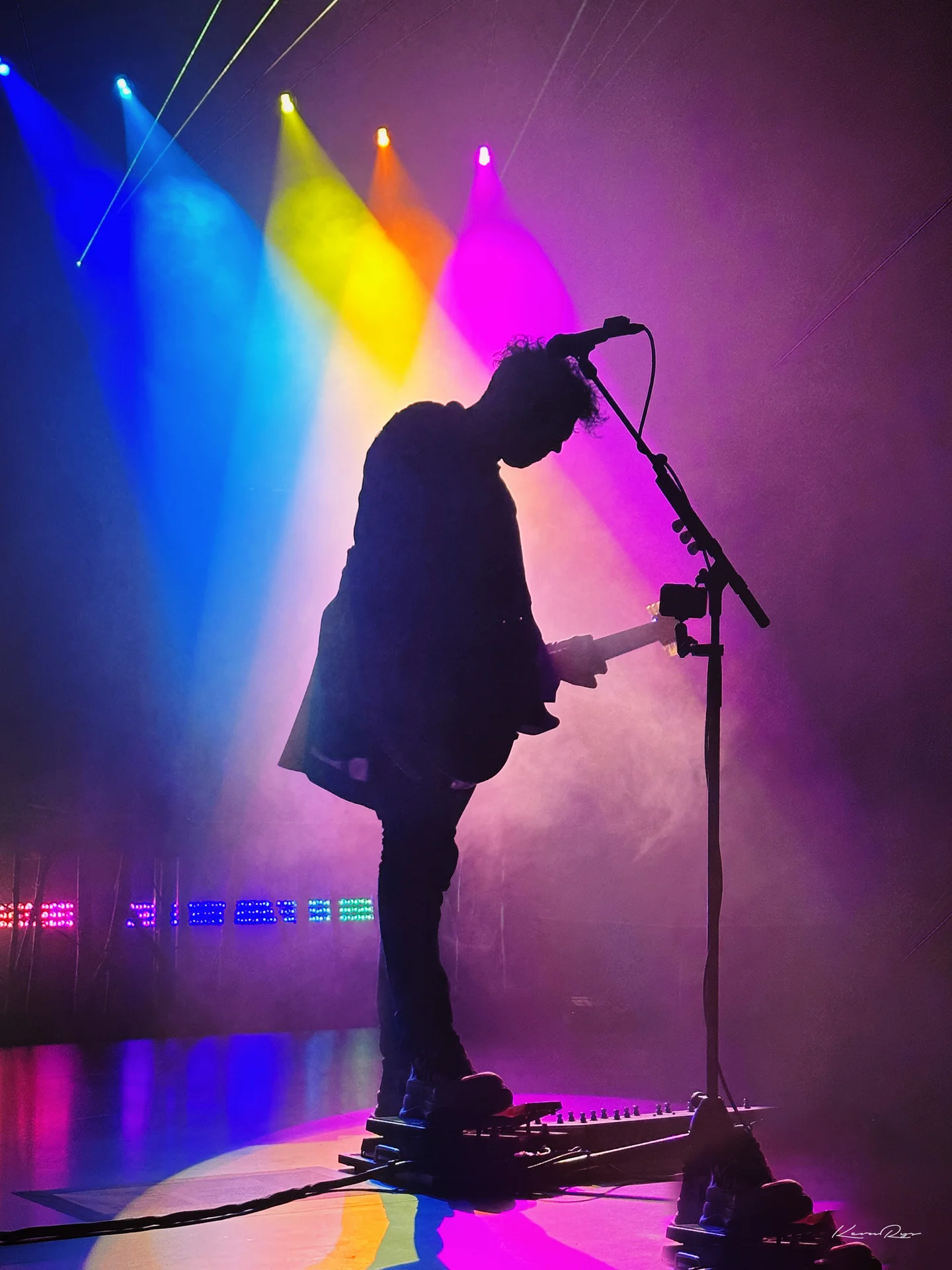 A guitarist in silhouette bows at the mic, backlit by blue, yellow, orange plus pink spotlights cutting through haze