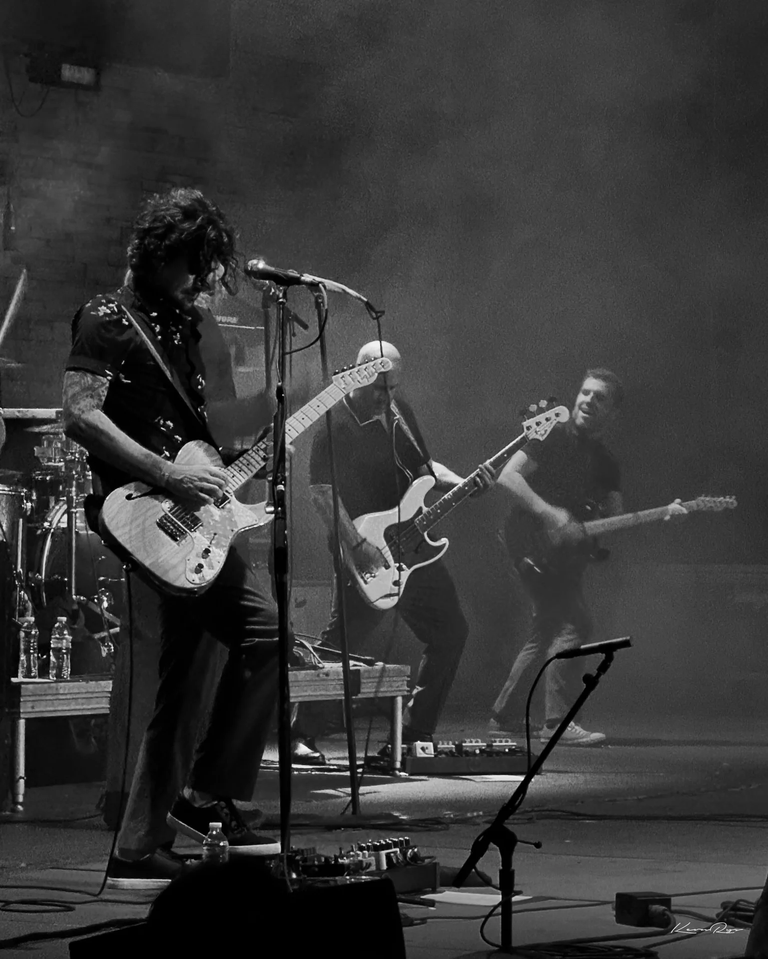 Three guitarists play through stage smoke in black-and-white, the nearest one at the mic with a drum kit behind
