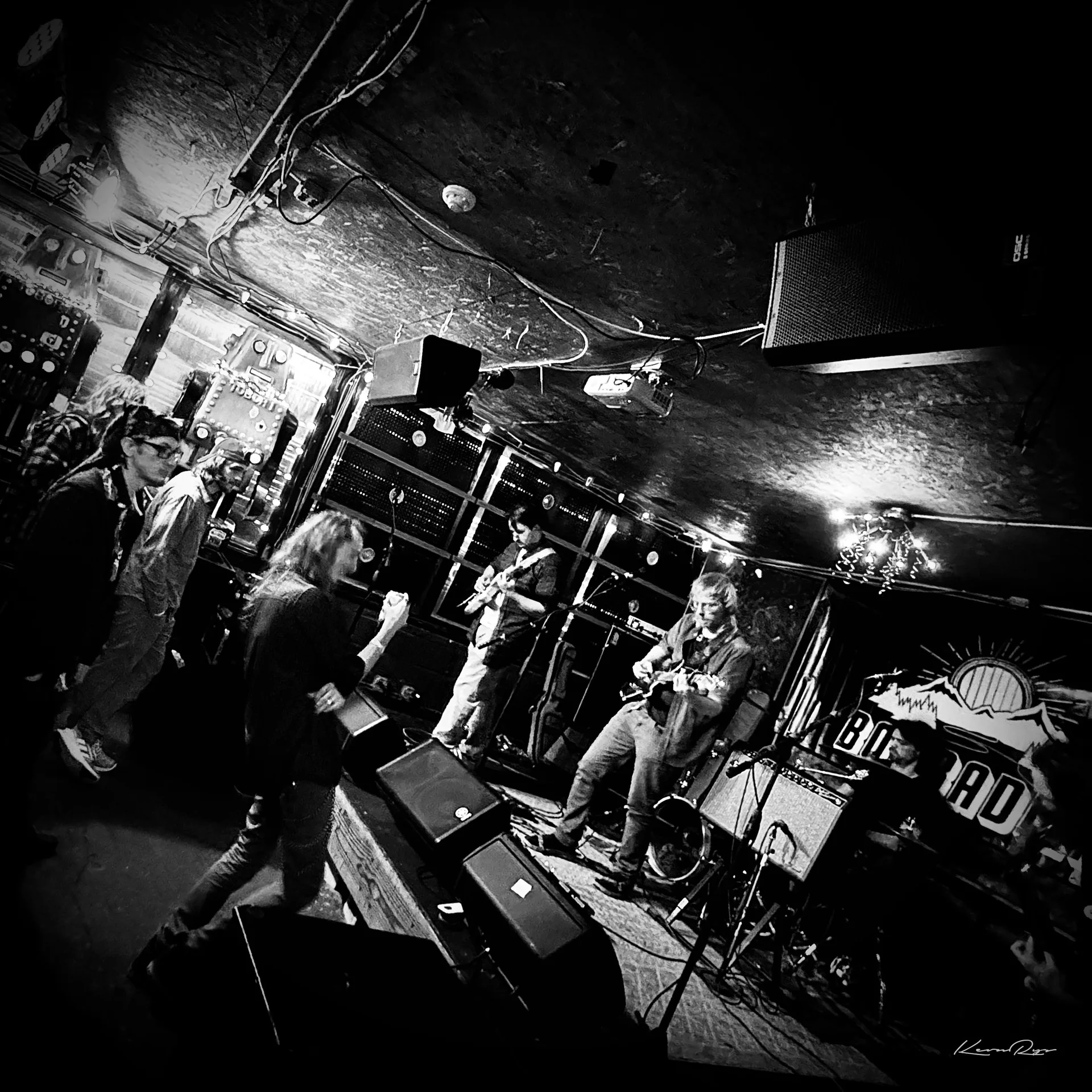 Tilted black-and-white view of a band in a cramped low-ceilinged room, speakers overhead plus fans dancing up front