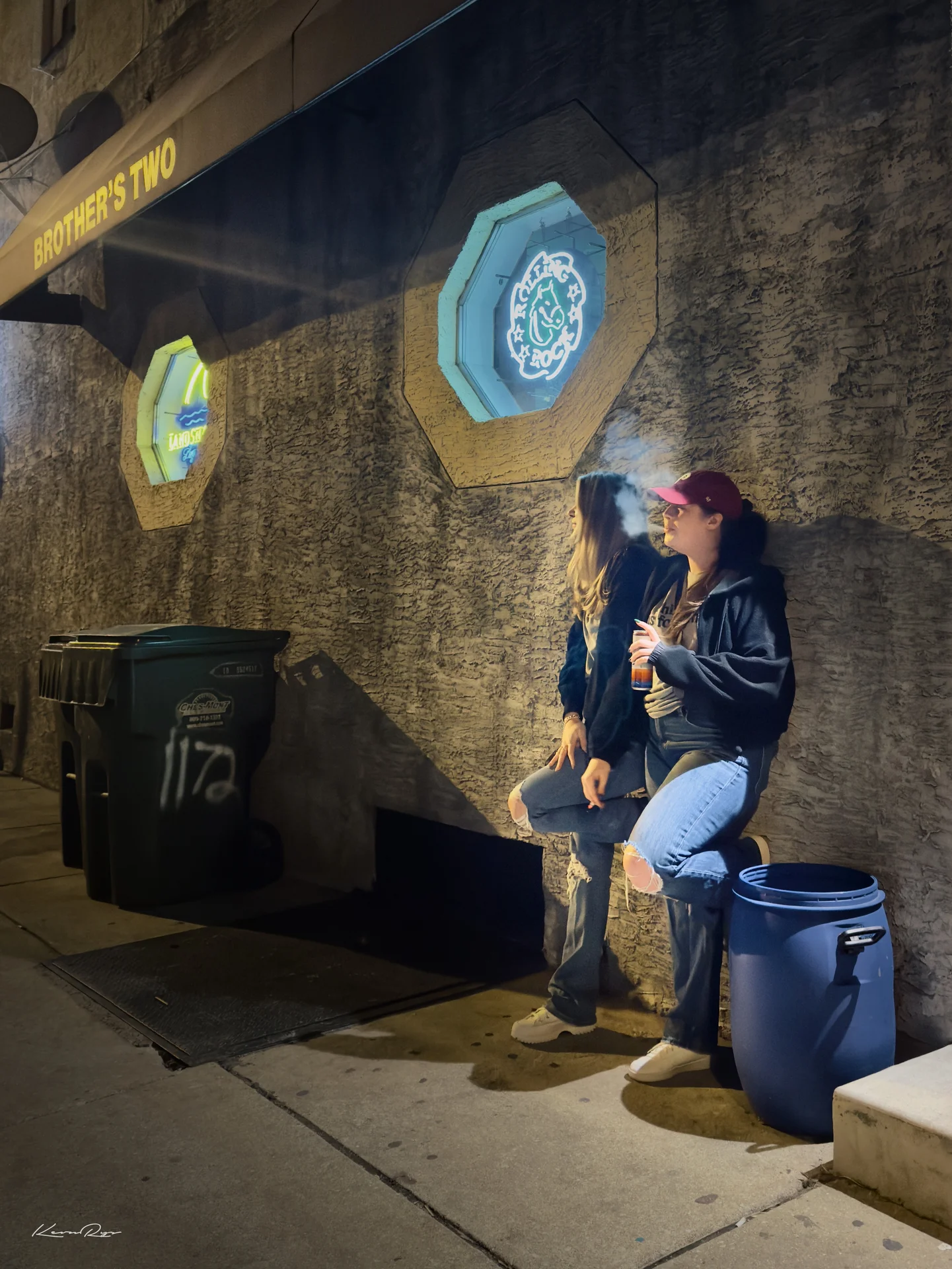 Two people lean on a stucco wall at night under the bar's awning, one in a red cap exhaling smoke beside neon window signs