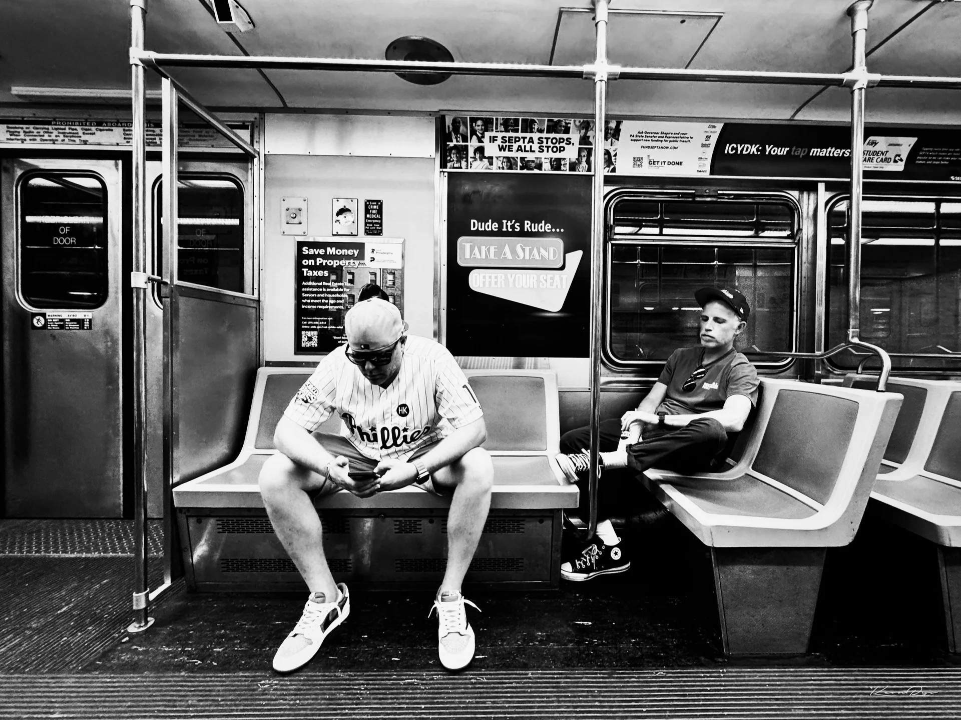 In black-and-white, a rider in a Phillies jersey checks a phone on a subway bench, beside an ad urging riders to offer their seat