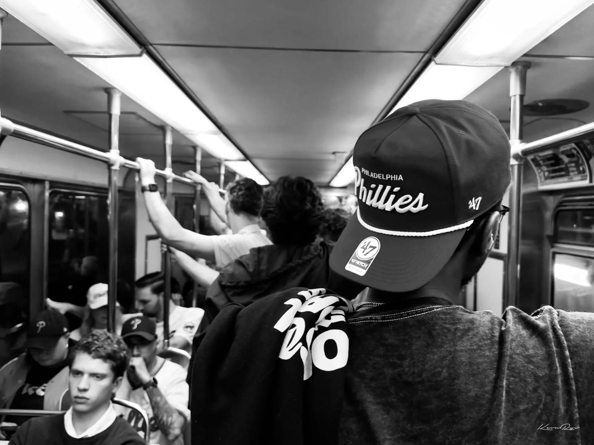 Black-and-white view over a fan's shoulder in a Phillies cap, looking down a crowded train car of riders holding the rails