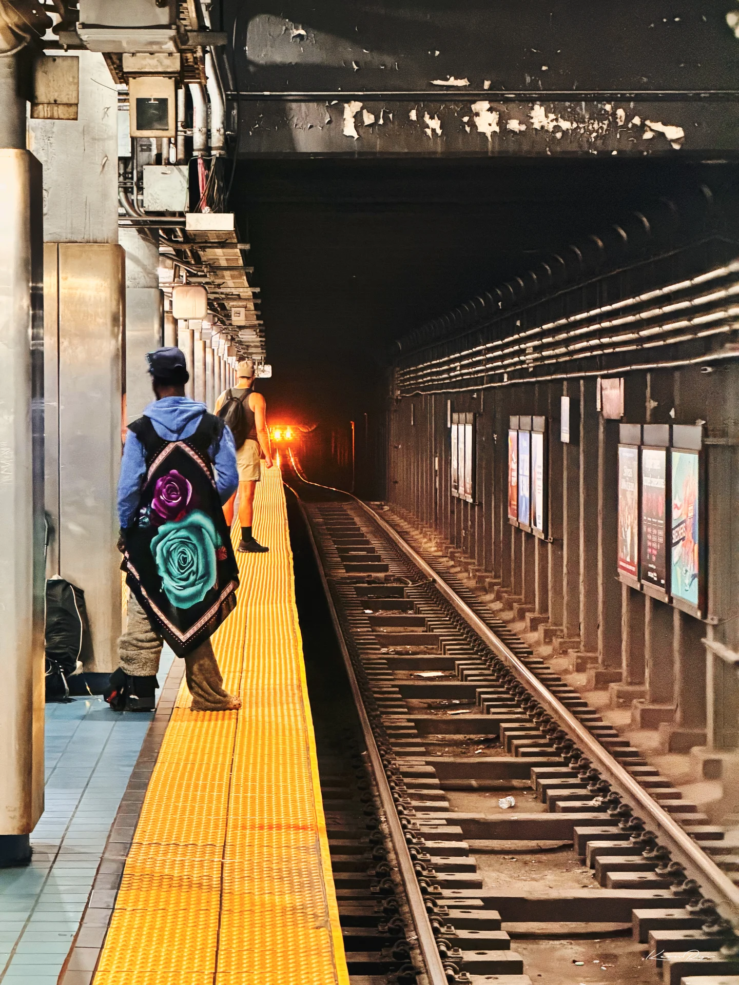 A rider in a rose-print shawl waits on the yellow platform edge as a train's red headlight glows far down the dark tunnel