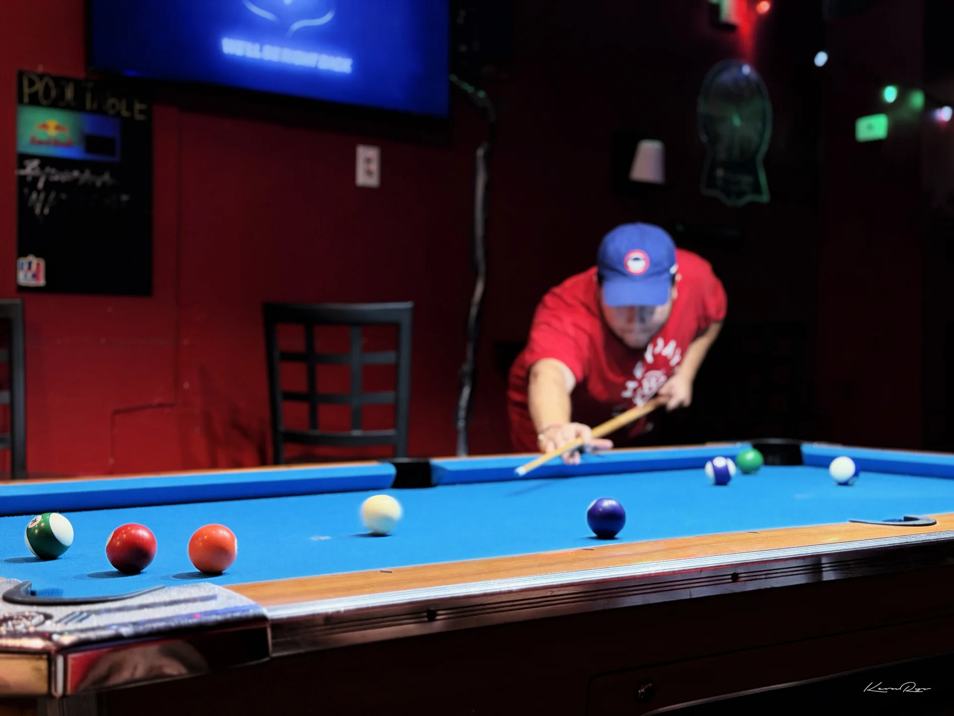 A player in a red shirt plus blue cap leans in to shoot across a blue felt pool table, balls scattered in the foreground