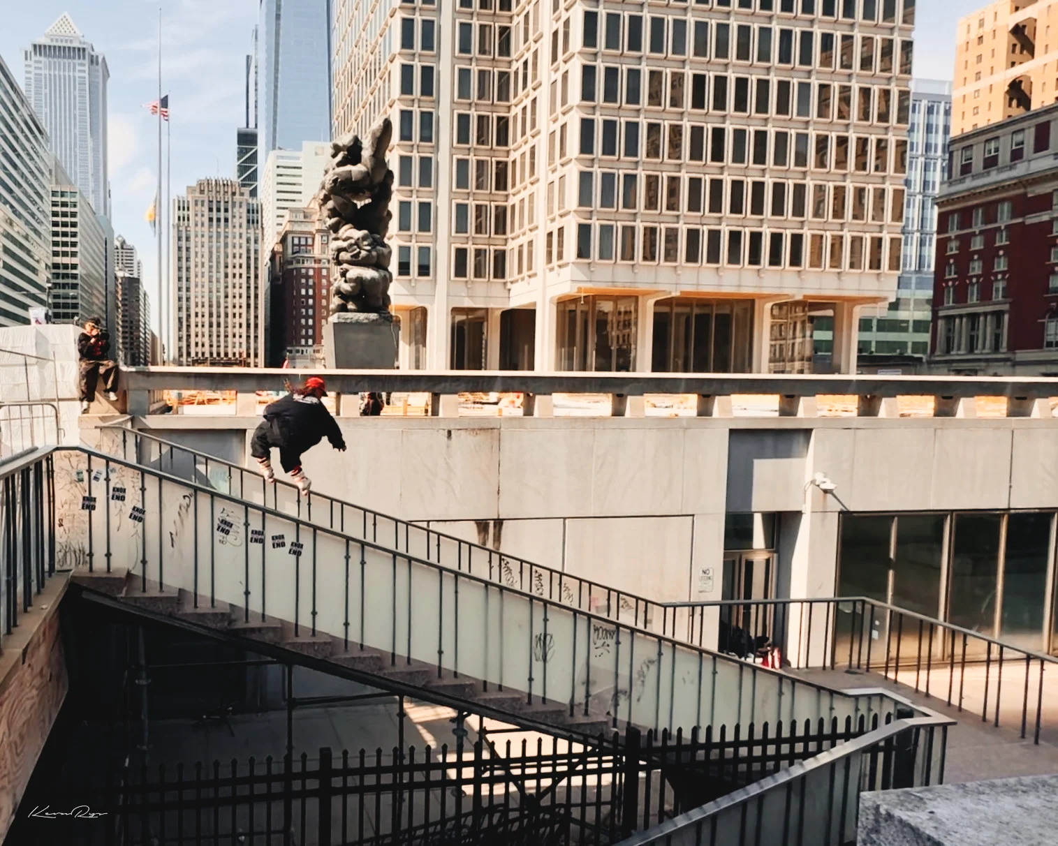 A person in a black jacket and red cap jumps along a stair railing beside a plaza, office towers rising behind