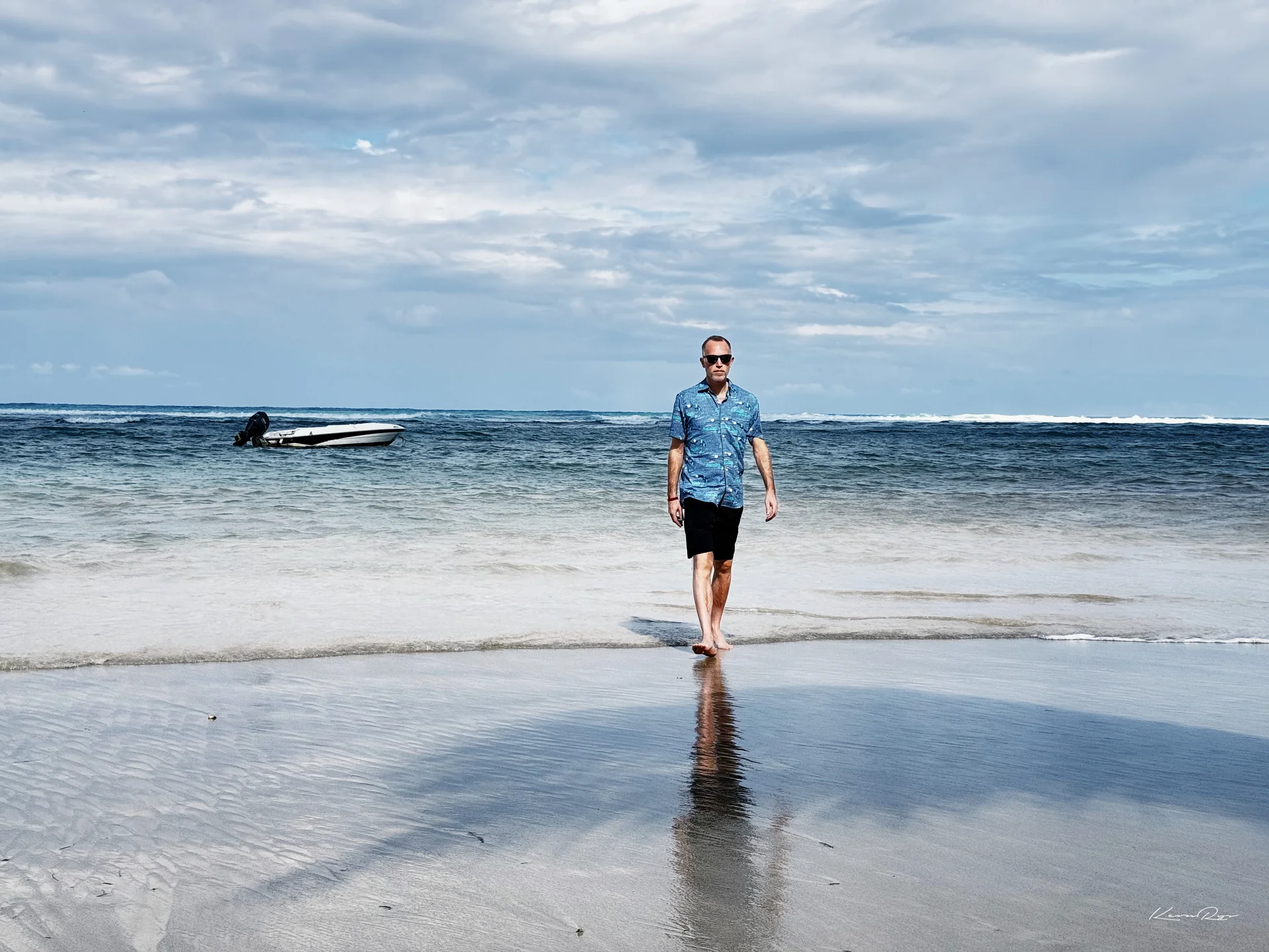 A person in a blue shirt walks barefoot out of the surf toward the camera, a small boat anchored behind under a cloudy sky