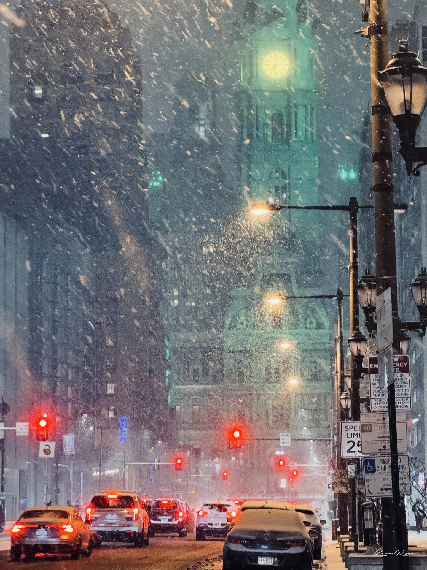 Snow falls thick over a street of cars stopped at red lights, City Hall's lit clock tower glowing green at the far end
