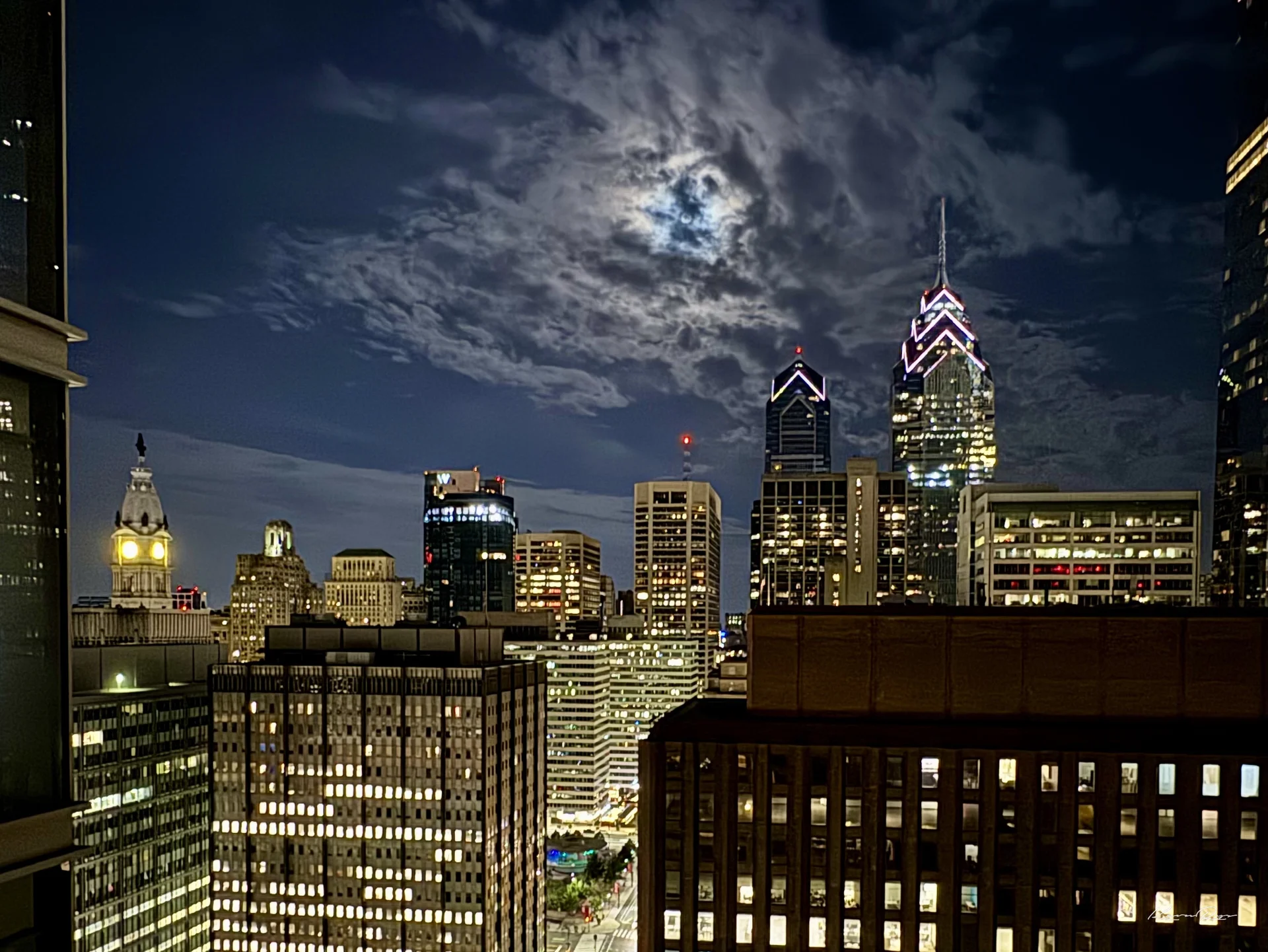 A full moon glows through clouds above lit office towers, City Hall's tower on the left plus the Liberty Place spires on the right
