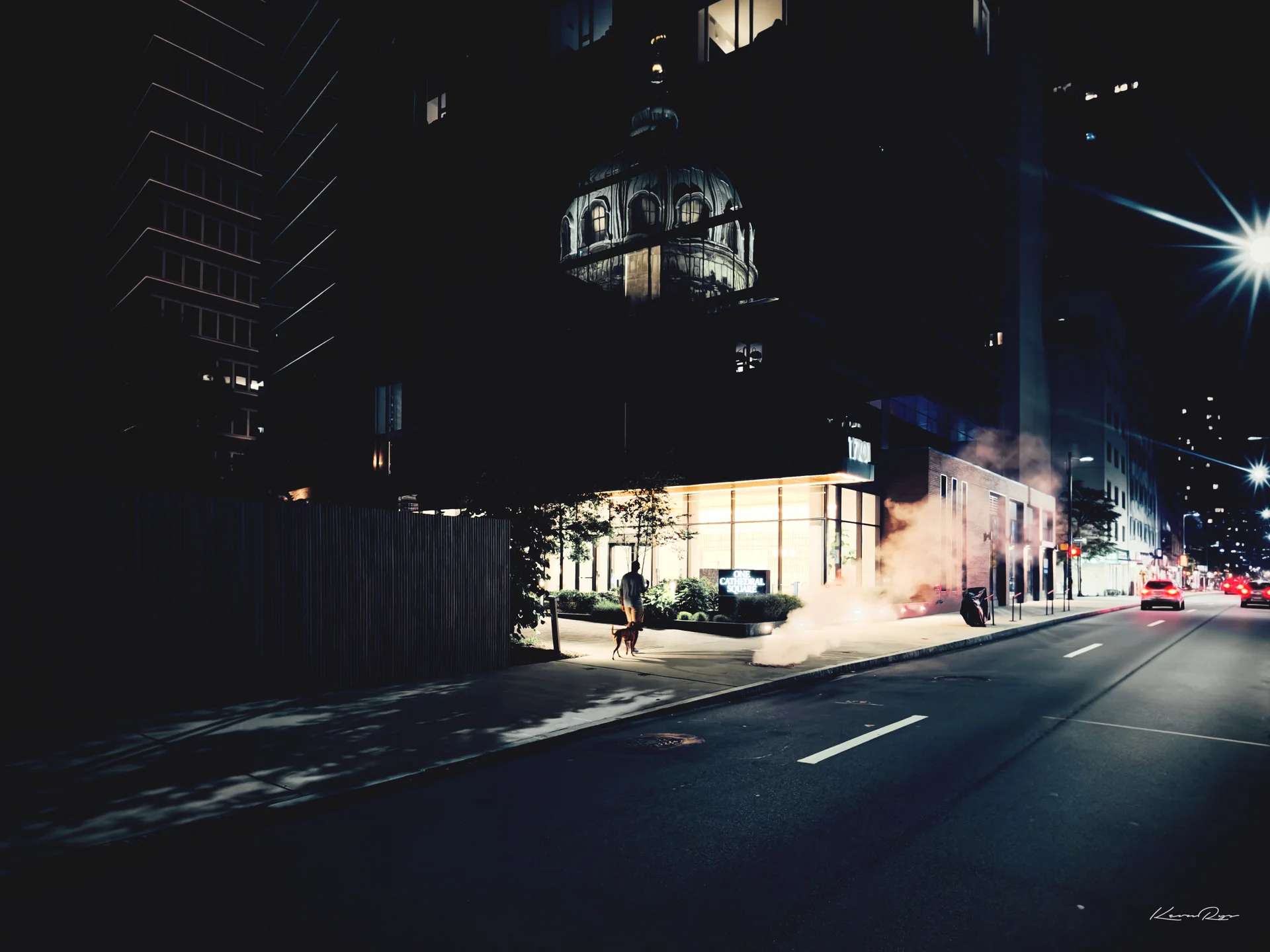 In color at night, a person walks a dog past a steam plume, a dome reflected in the dark glass tower above, streetlights flaring