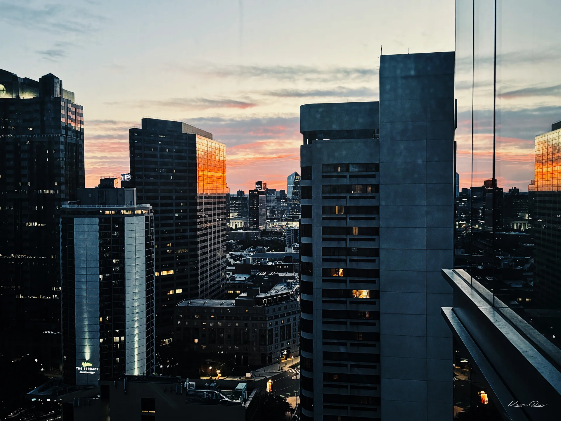 Orange sunset light reflects off glass towers in silhouette, a dark high-rise in the foreground with a few windows lit