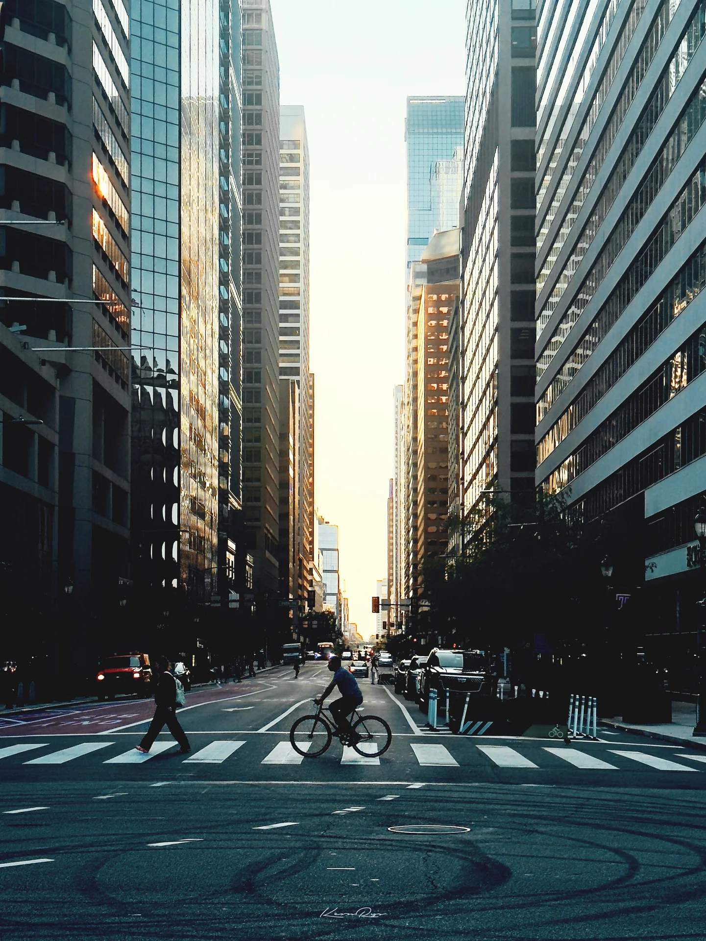 A cyclist plus a pedestrian cross at the crosswalk, tall office towers framing a hazy glow at the far end of the street