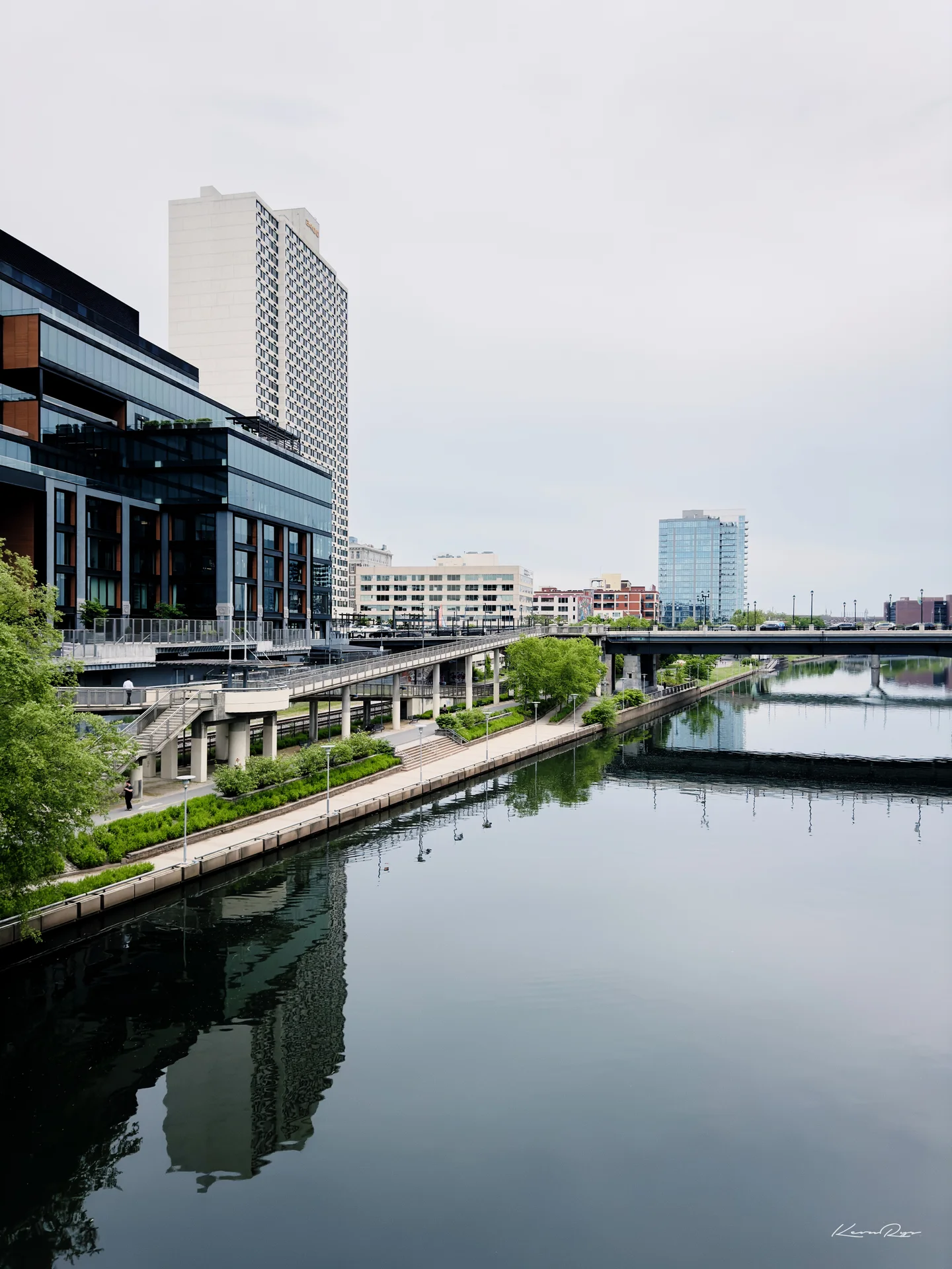 A calm river reflects glass towers, a riverside walkway lined with plants, plus a bridge under a gray sky