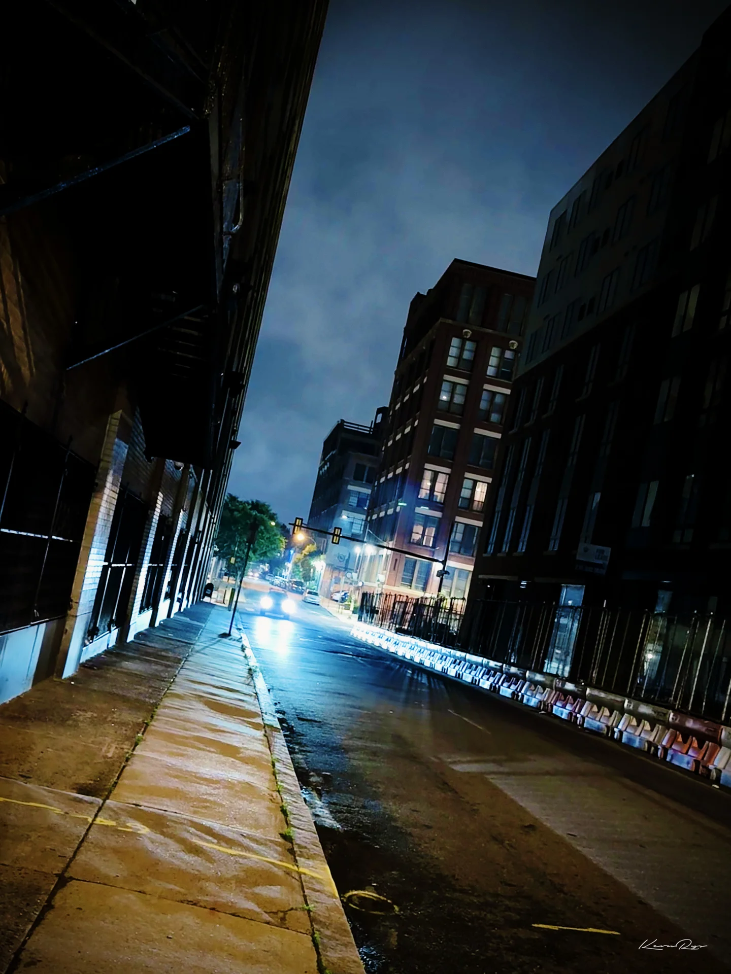 A tilted view of a wet, empty street at night, with brick buildings, traffic lights plus distant headlights