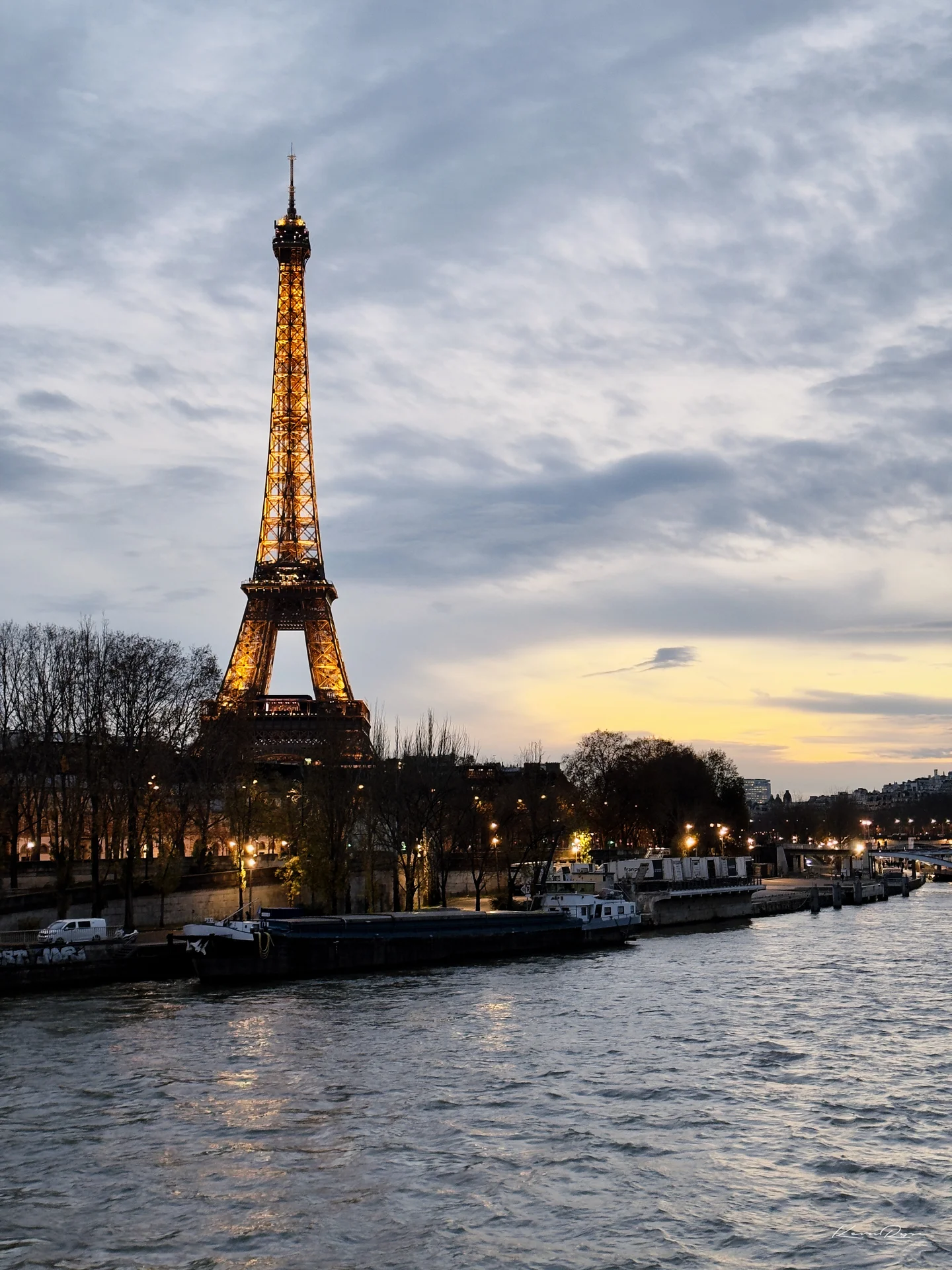 The lit Eiffel Tower glows gold under a cloudy sky, with barges moored along the dark river below