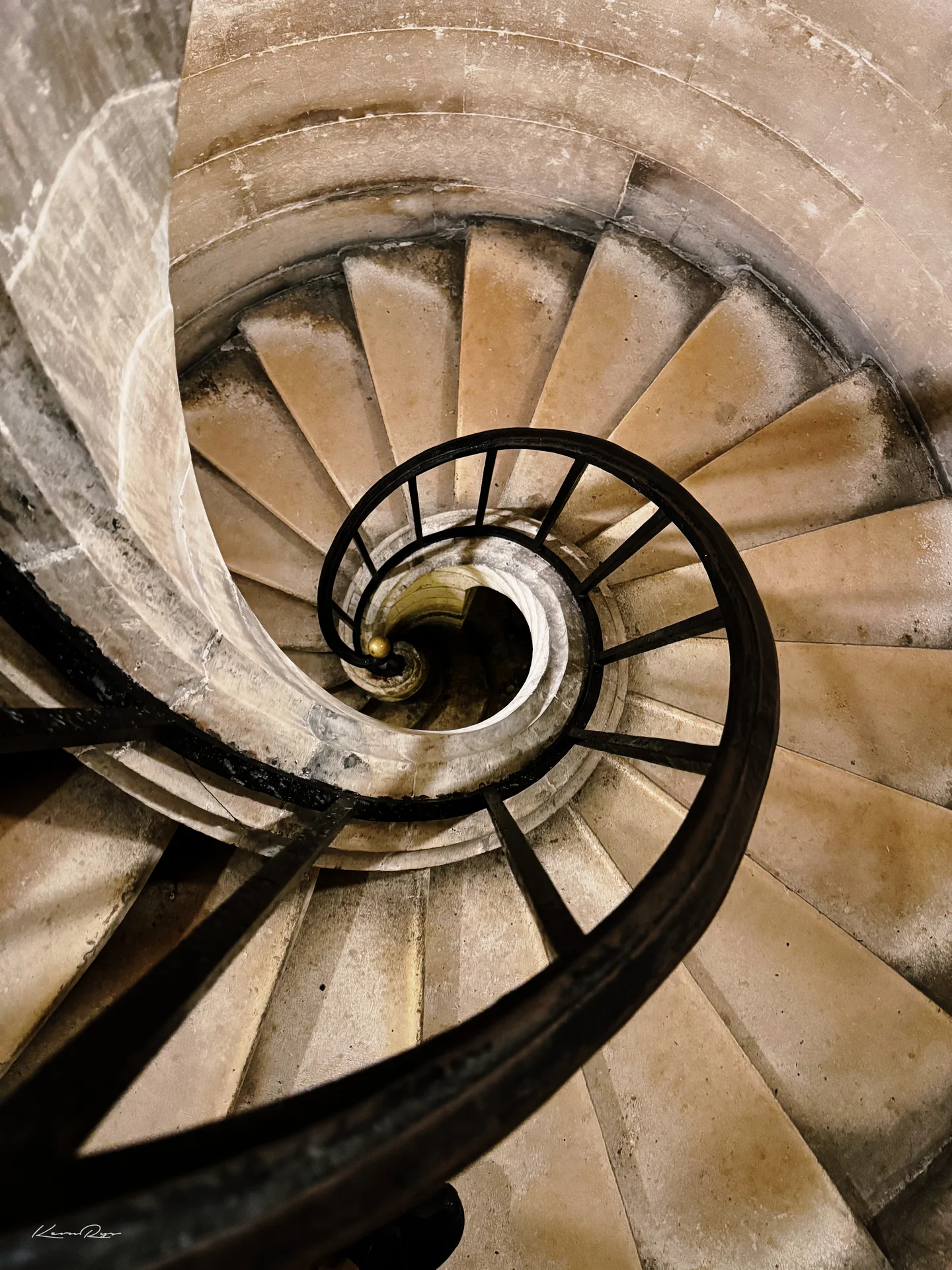 Looking down a stone spiral staircase, its dark iron railing curling toward the center