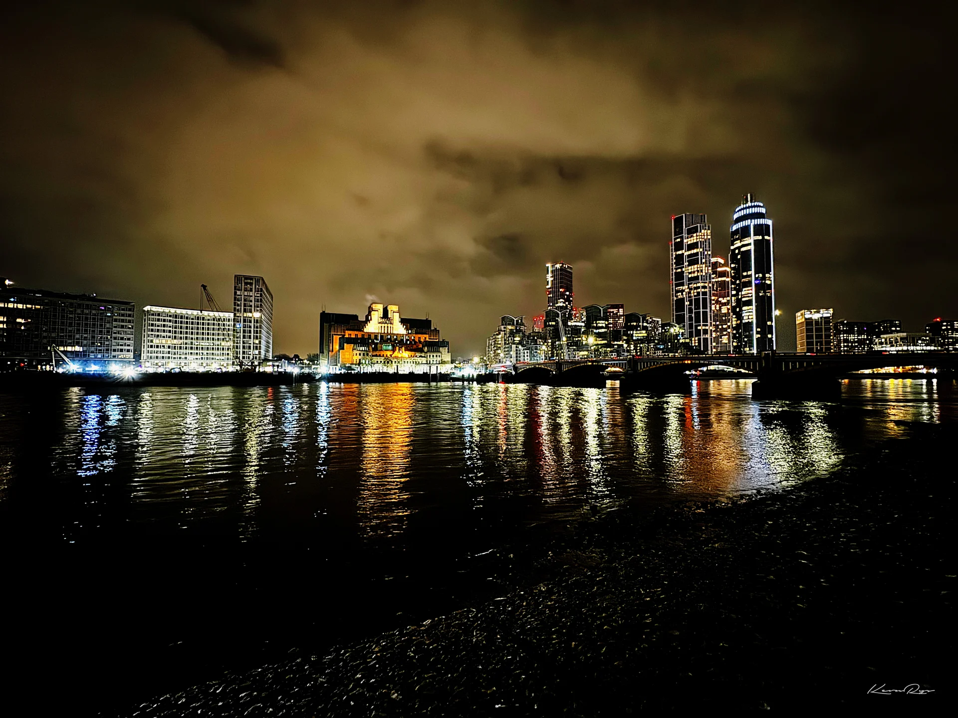 City lights and tall towers reflect in dark water at night, seen from a pebbly shore under orange clouds