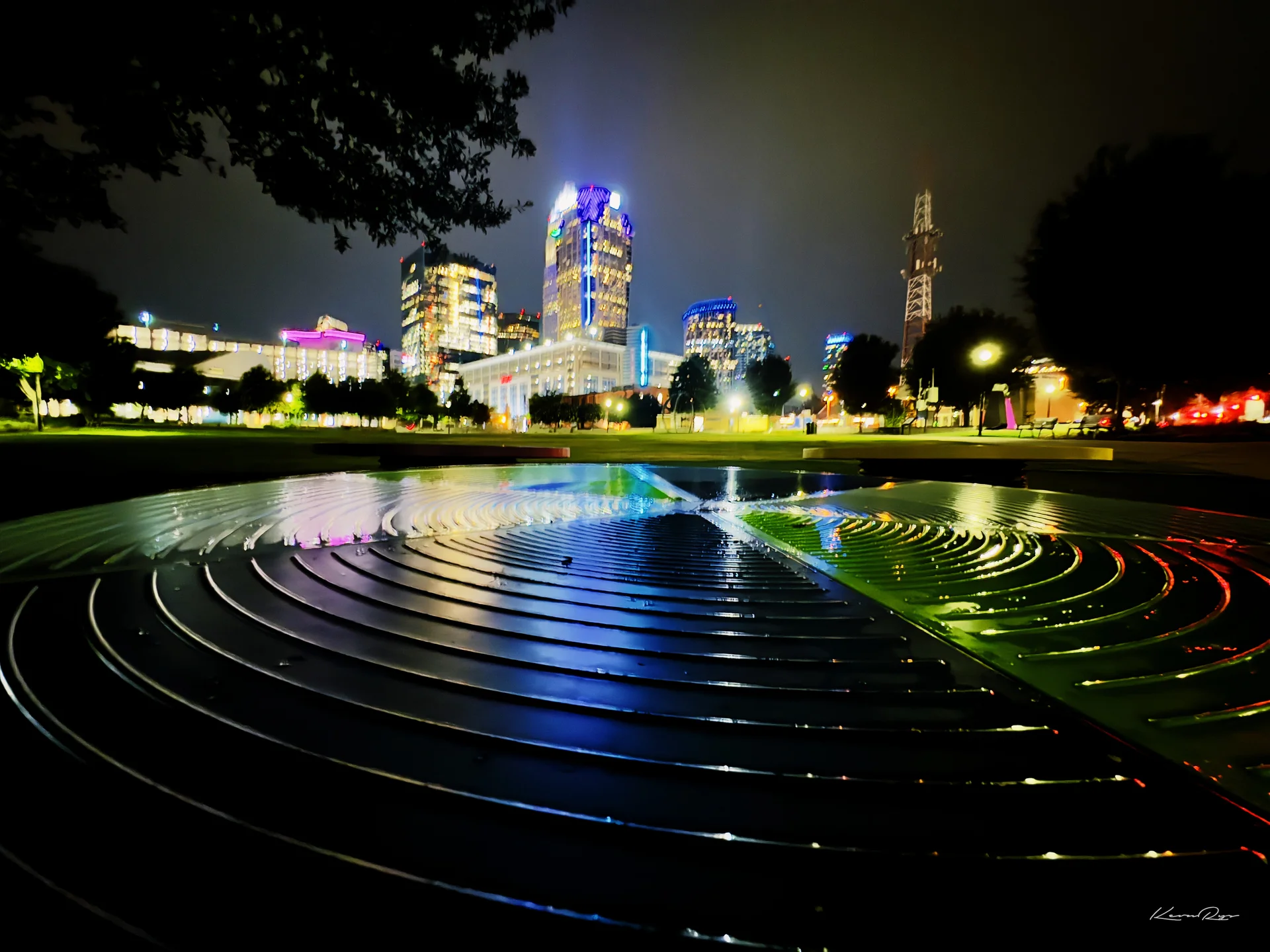 A lit skyline at night behind a wet, curved metal sculpture glowing with blue, green plus red reflections