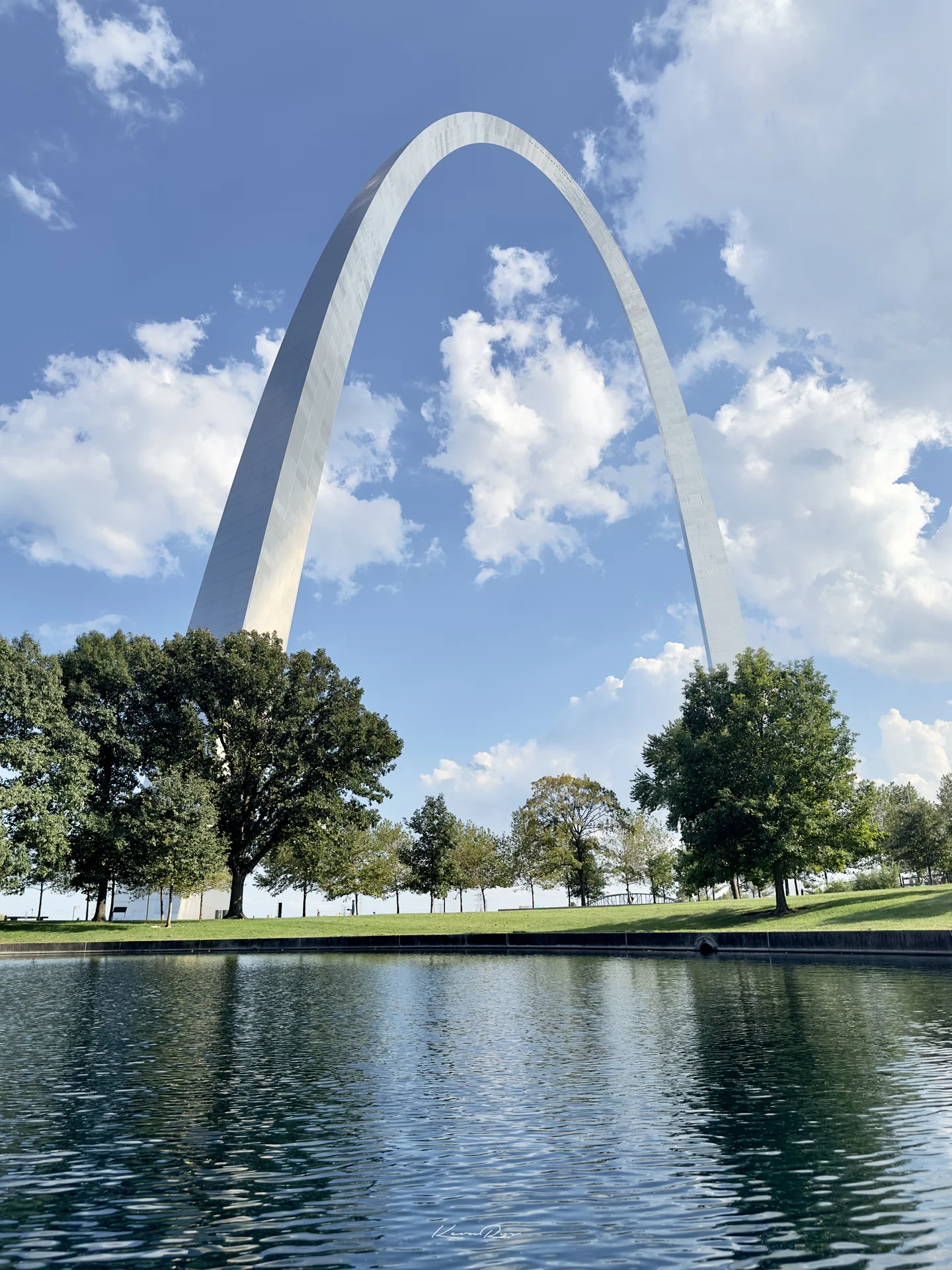 The silver arch rises over green trees under a blue sky with white clouds, mirrored in a pond