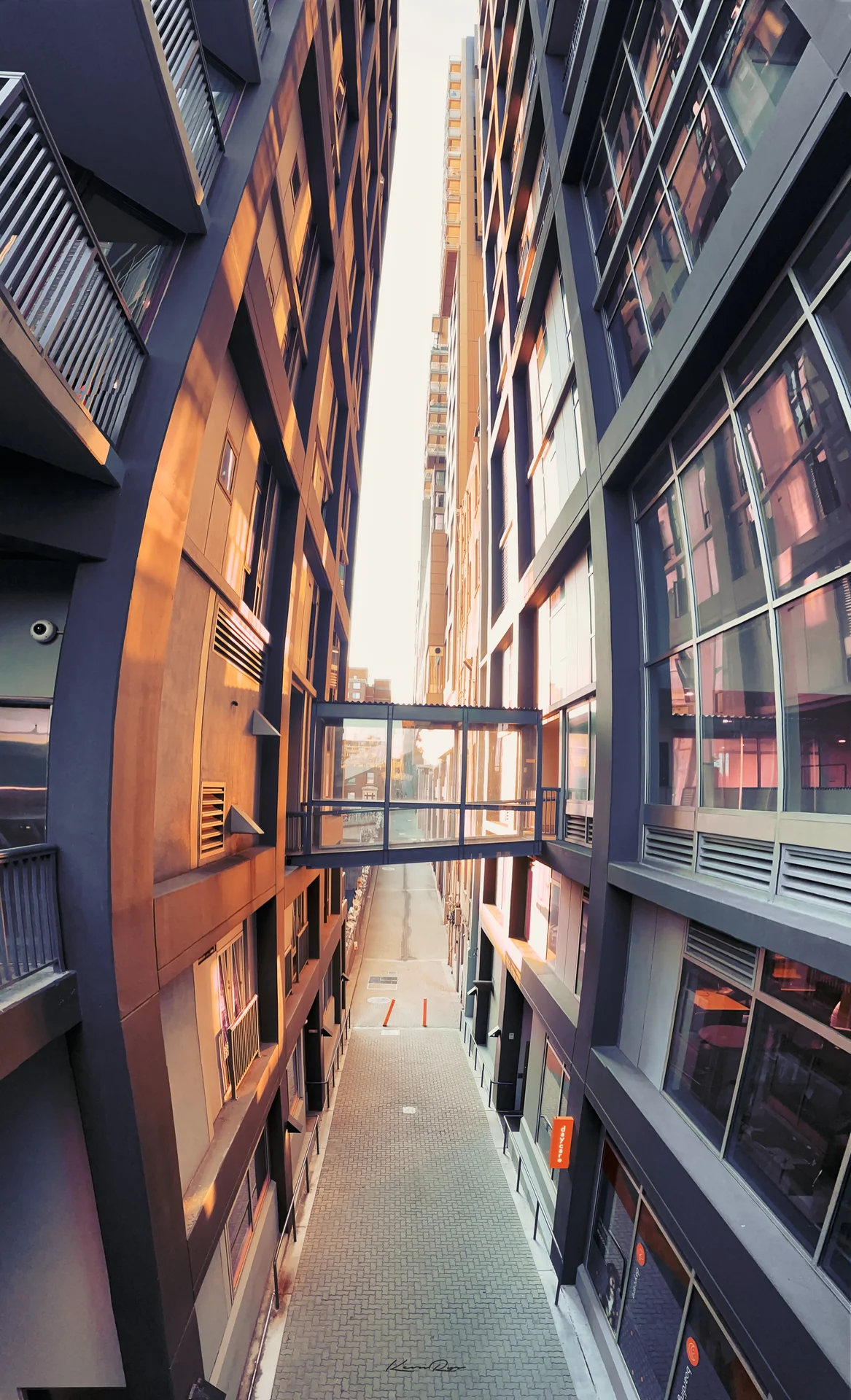 A glass walkway spans a narrow brick alley between tall buildings, warm light on one wall