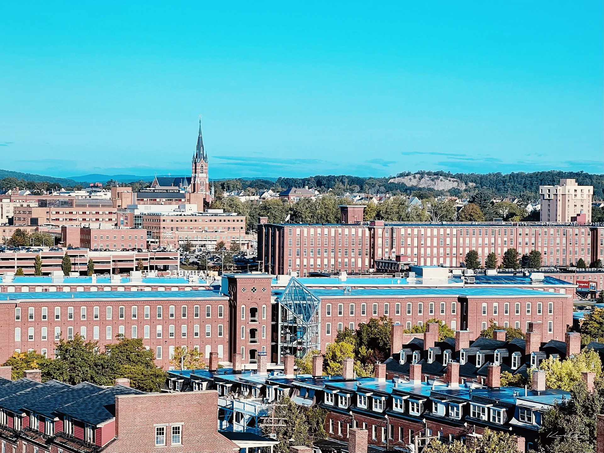Rows of red brick mill buildings under a clear blue sky, with a church spire plus wooded hills behind