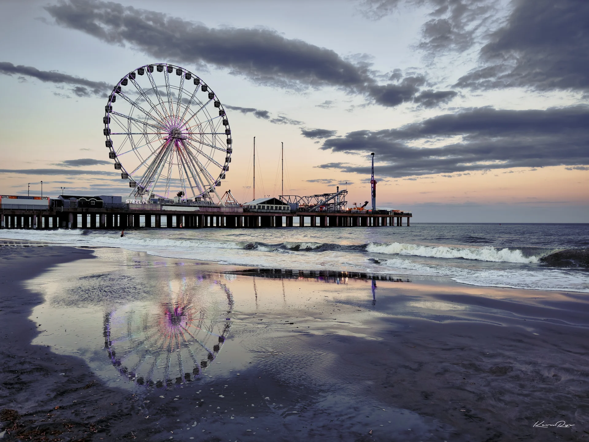 A Ferris wheel on a pier at dusk, reflected in wet sand as waves roll onto the beach