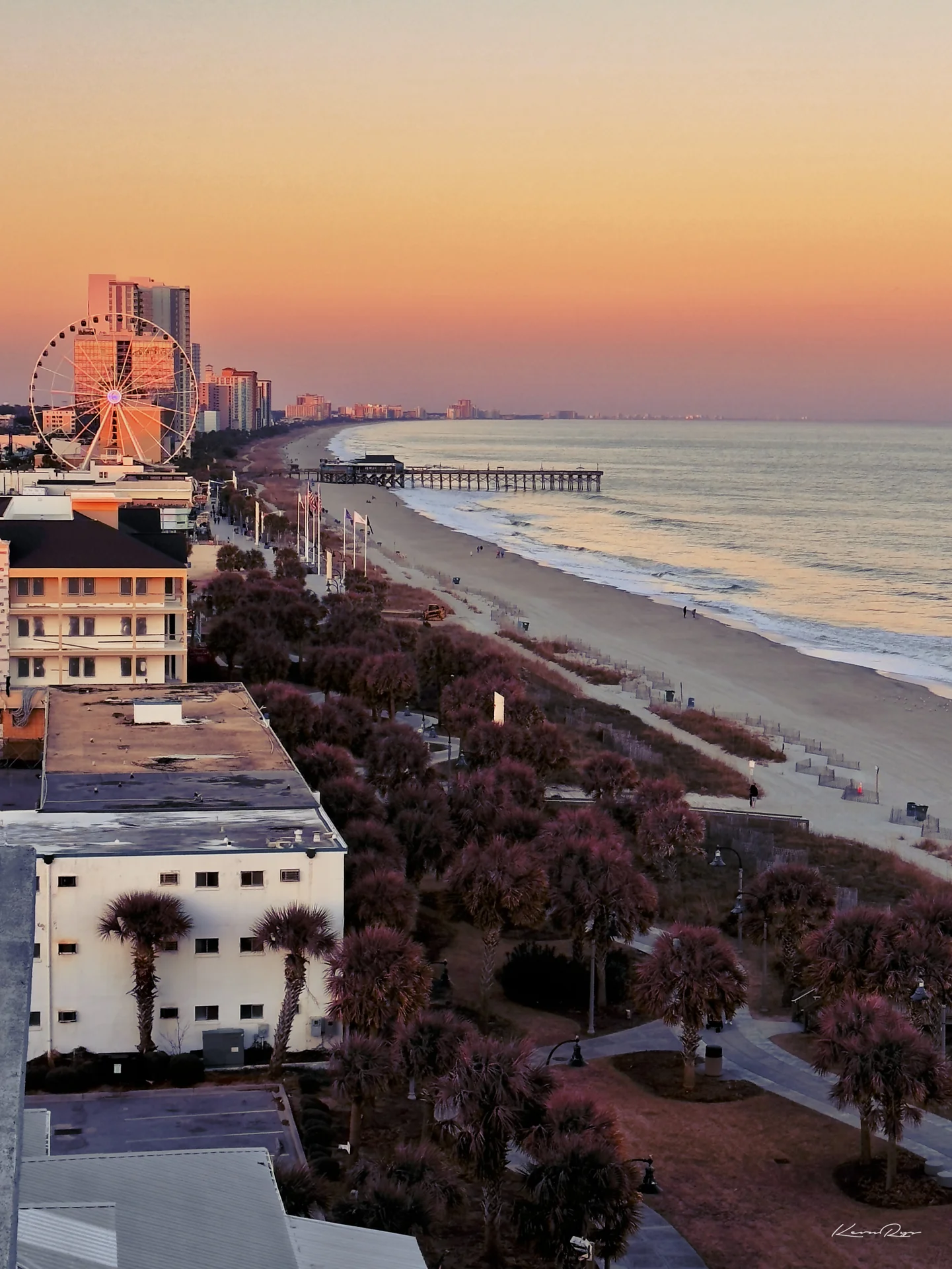 An orange sunset over a long beach seen from above, with a Ferris wheel, a fishing pier plus palm trees