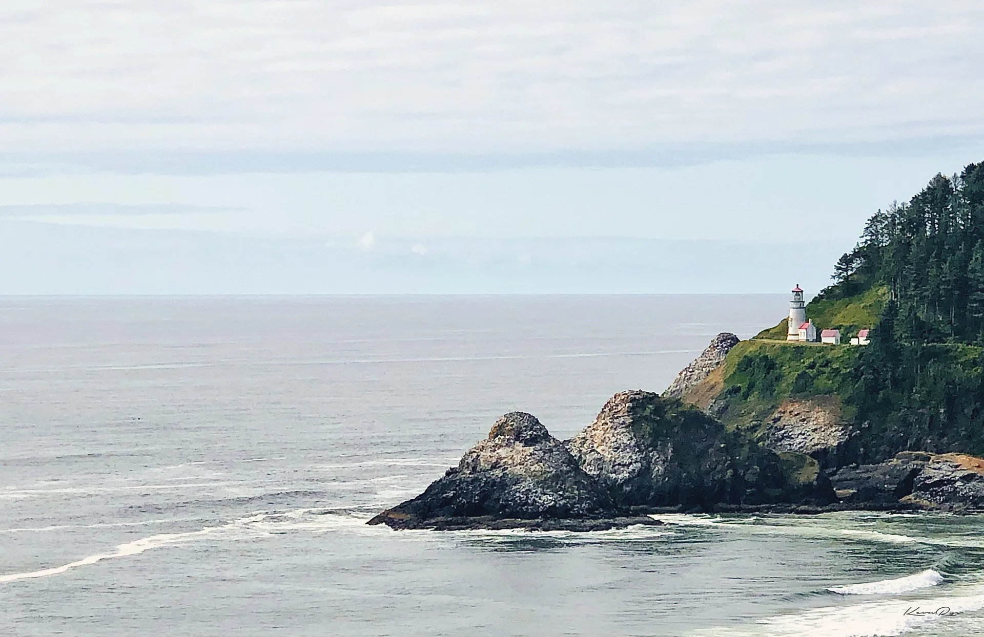 A white lighthouse sits on a green, forested headland above rocky outcrops in a gray sea