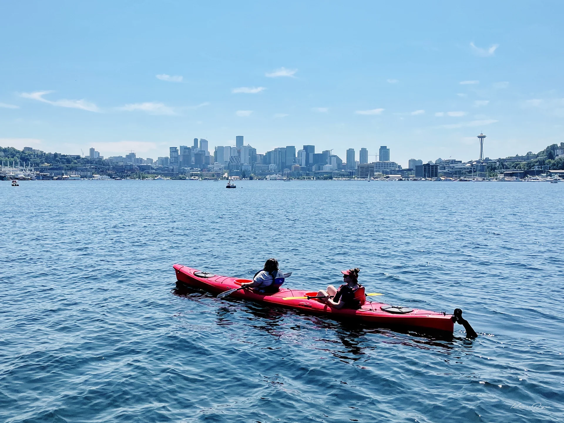 Two people paddle a red kayak on blue water, with a city skyline under a clear sky behind them