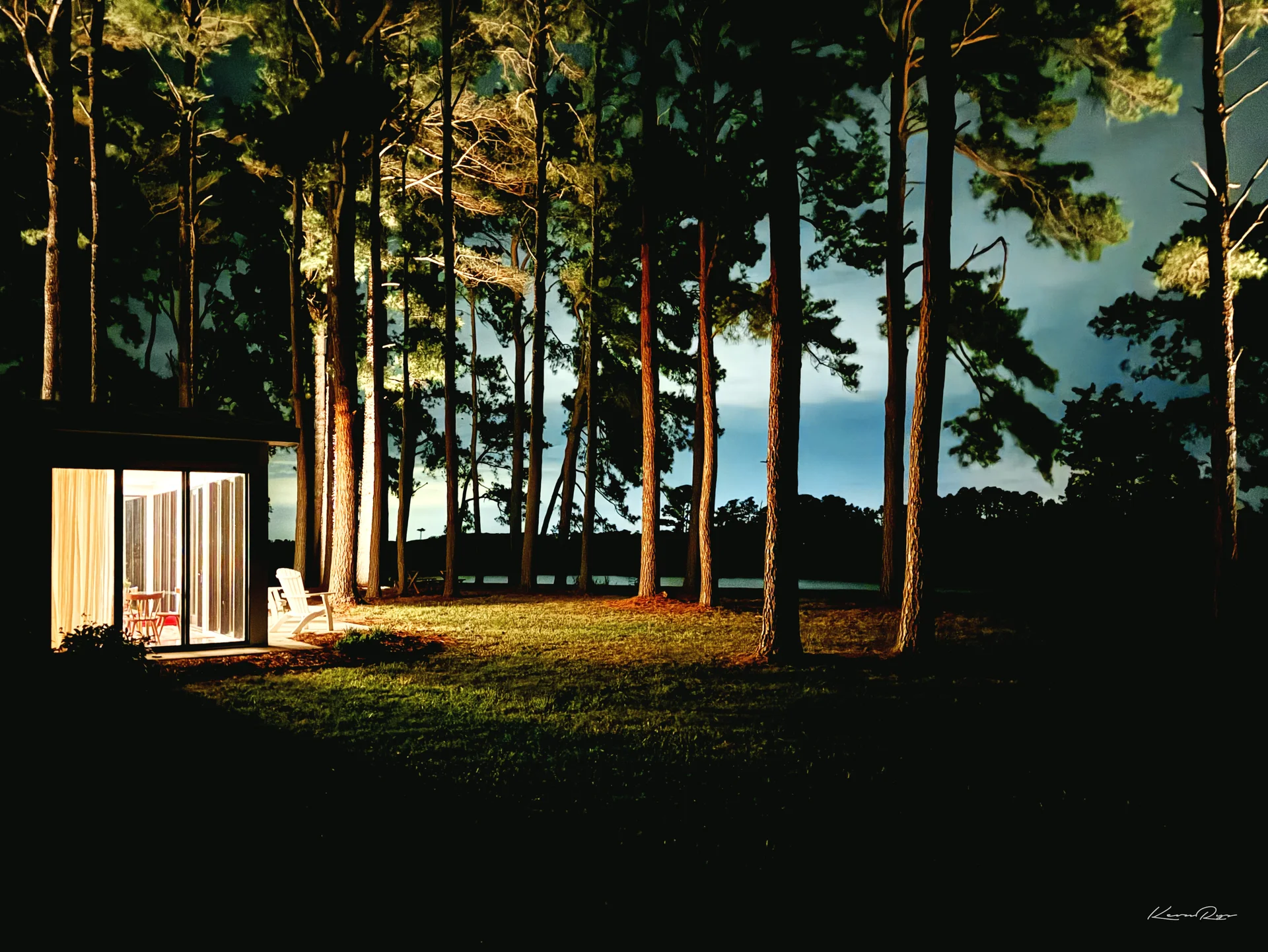 A small glass-walled cabin glows warm at night beside tall pines lit from below, a dark lawn plus water behind