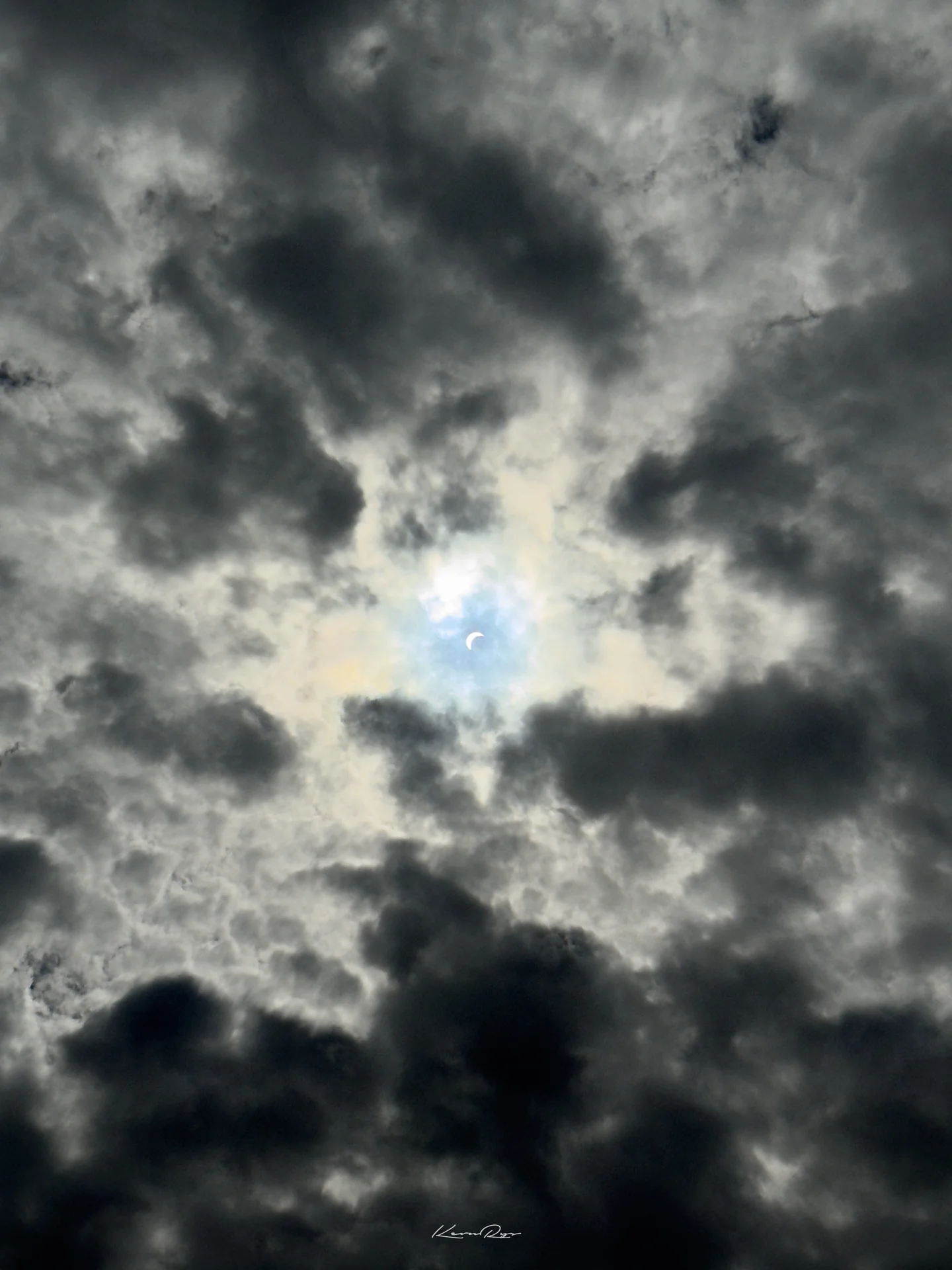 A thin crescent of sun peeks through a small gap in heavy gray clouds, a pale blue halo around it