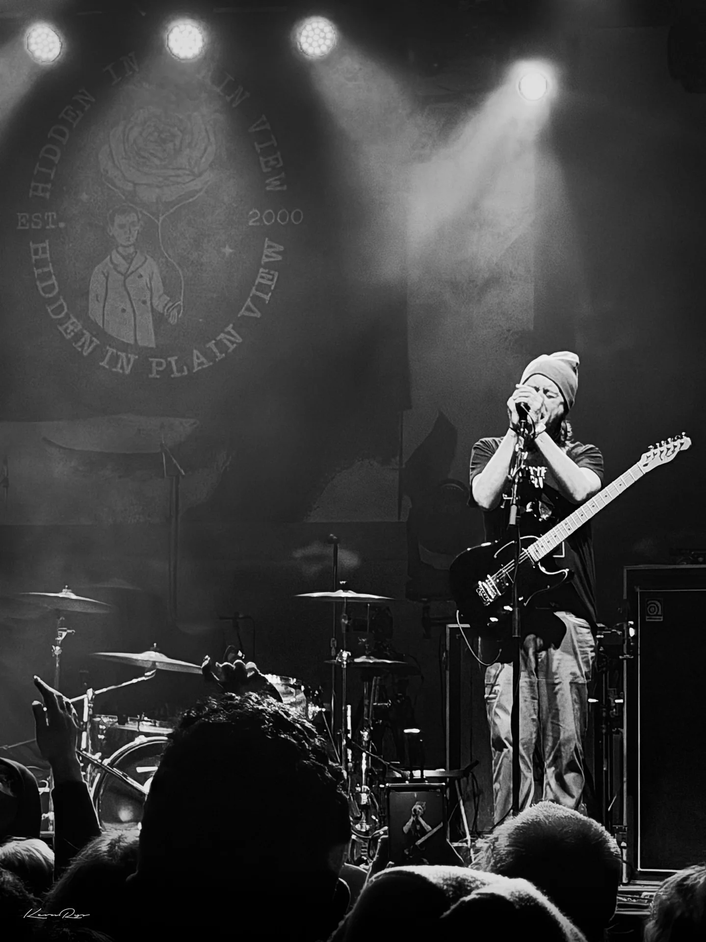 In black-and-white, a singer in a beanie cups the mic with a guitar slung low, fans' hands raised in front of the stage
