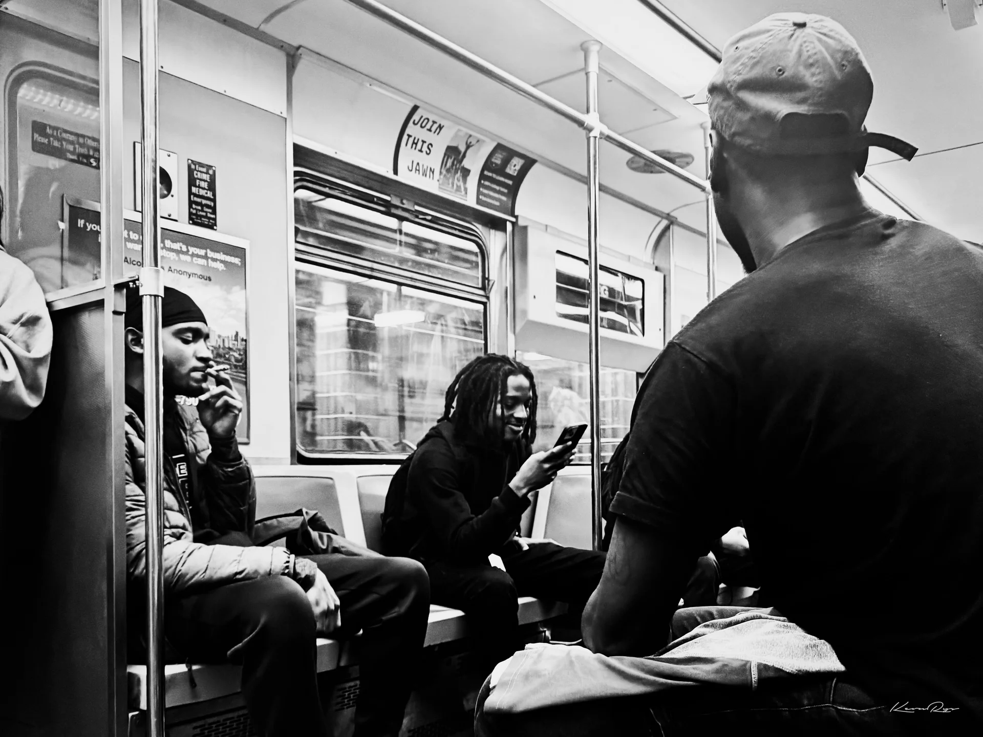 In black-and-white, riders sit in a subway car, one smoking, one smiling at a phone, another in a backward cap in front