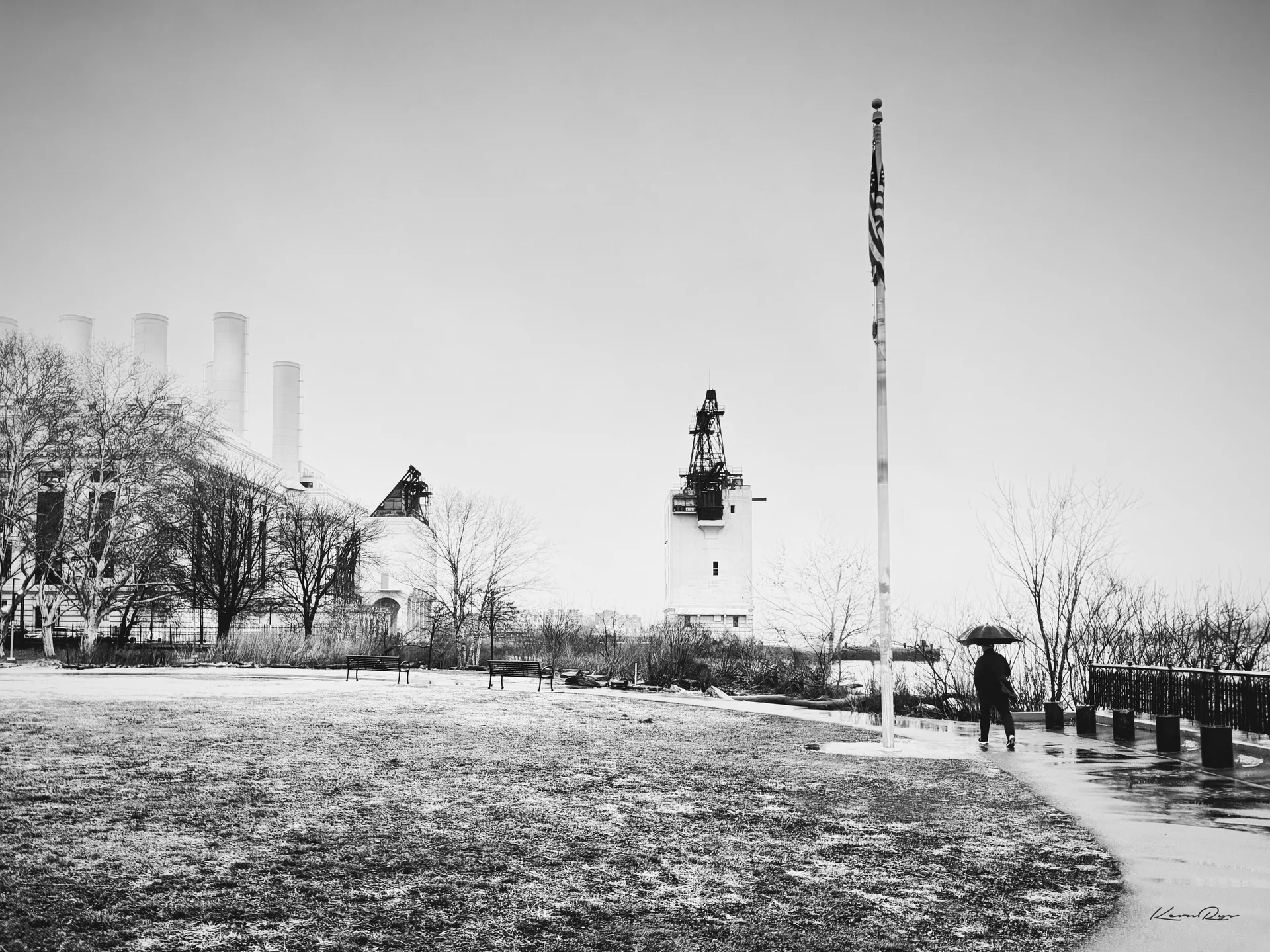 In black-and-white, a lone walker with an umbrella crosses a frosty lawn past a tall flagpole, smokestacks plus a crane tower in the mist