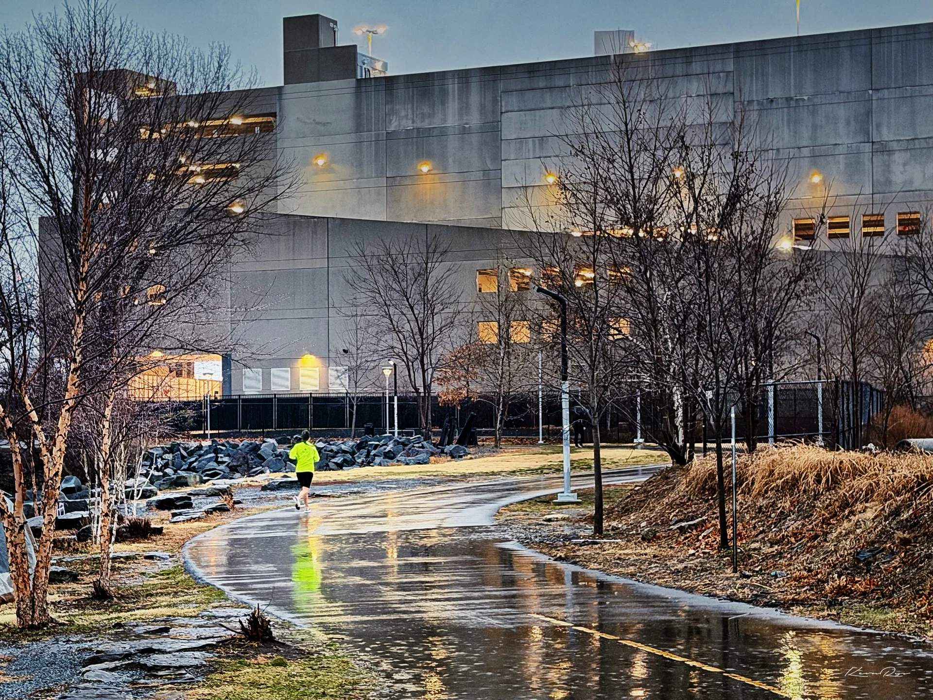 A runner in a neon yellow shirt jogs along a wet, curving path at dusk, lit windows of a large concrete building behind