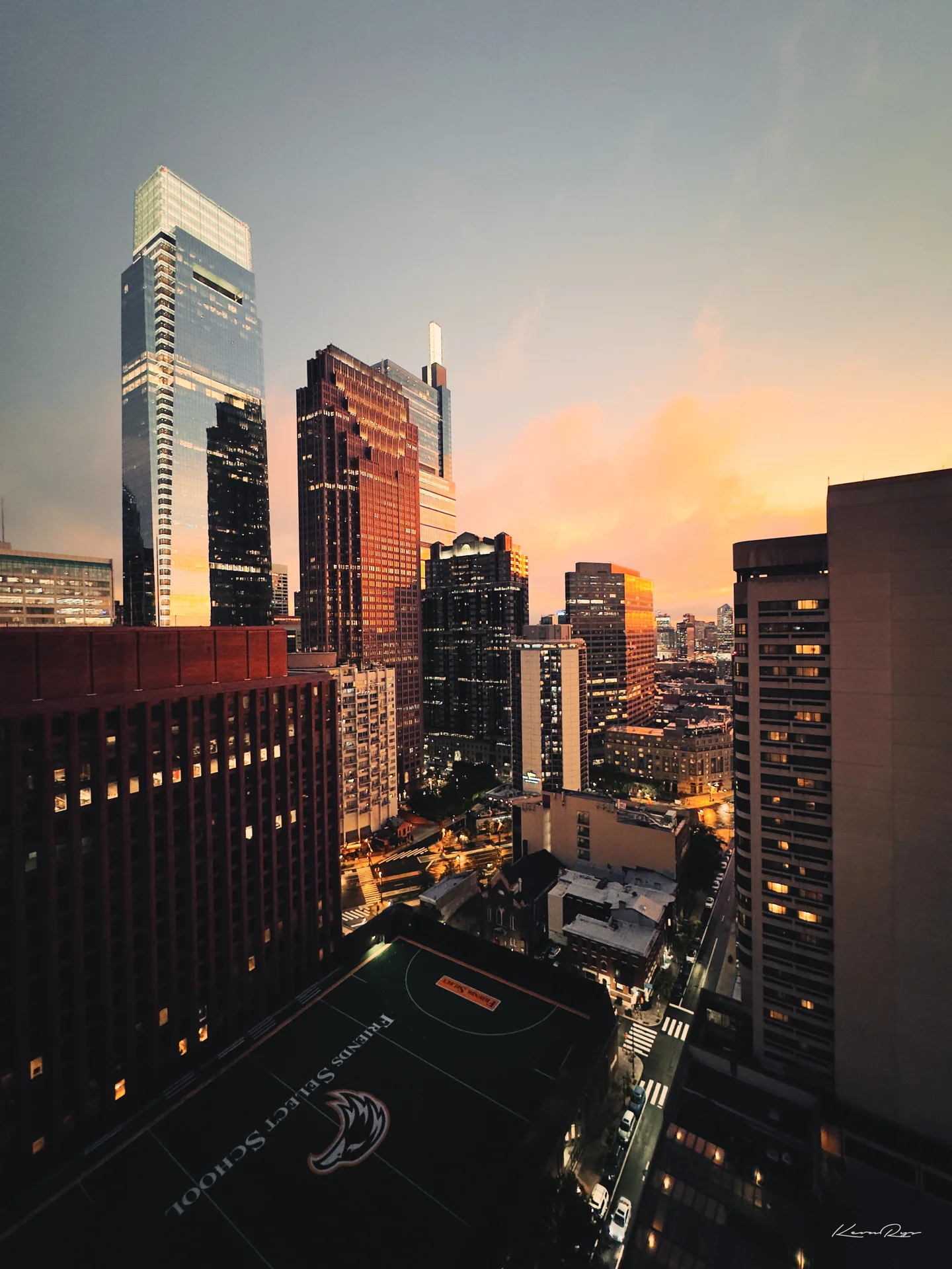Glass towers catch a pink sunset above rows of lit windows, a rooftop basketball court far below