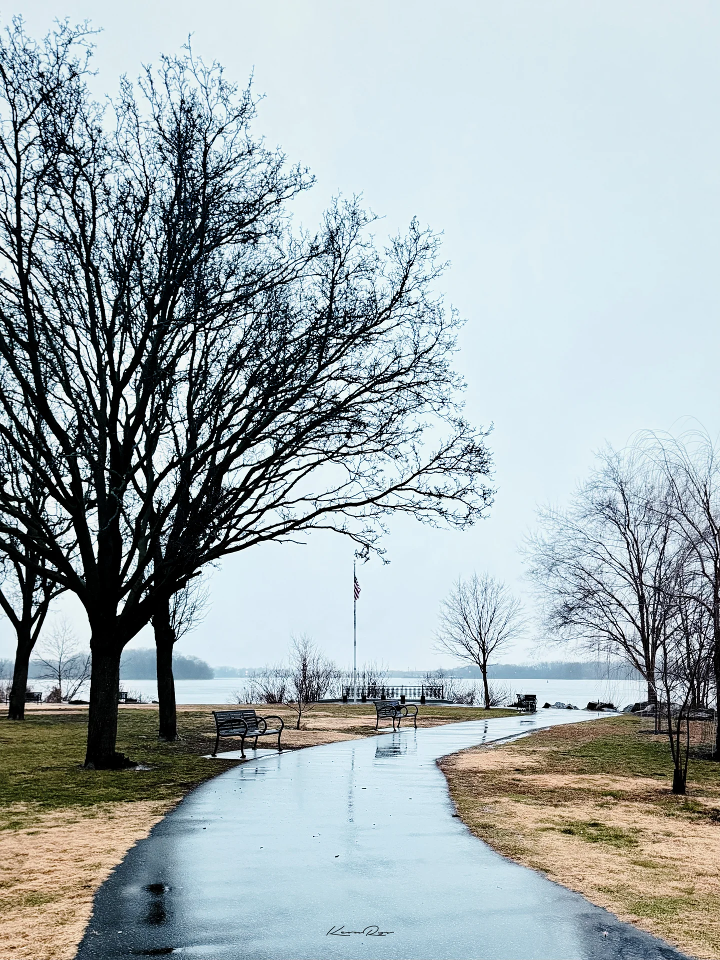 A wet path curves past bare trees plus empty benches toward a flagpole, the river gray under an overcast sky