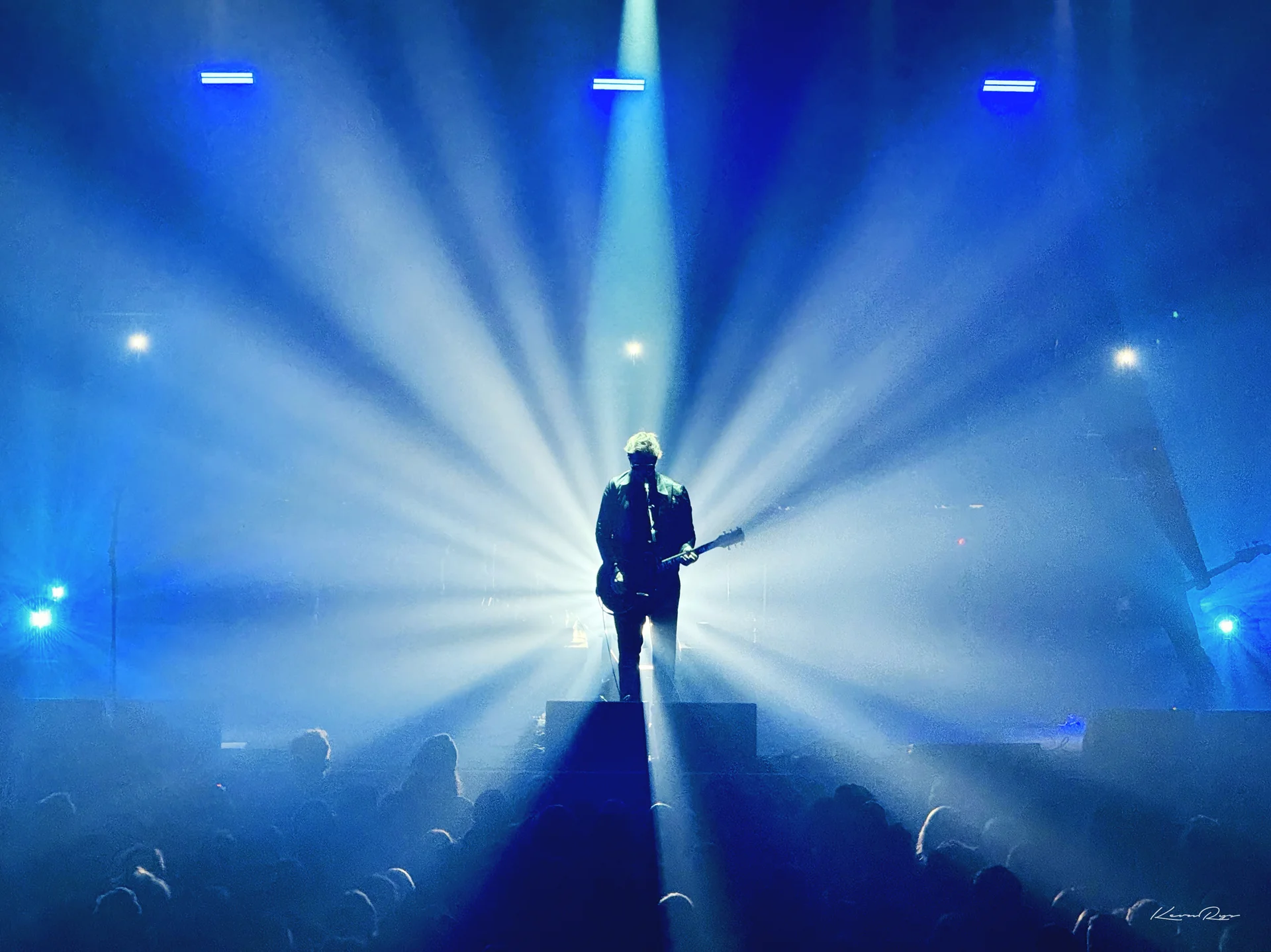 A singer with a guitar stands silhouetted at center stage, blue light beams fanning out over the crowd
