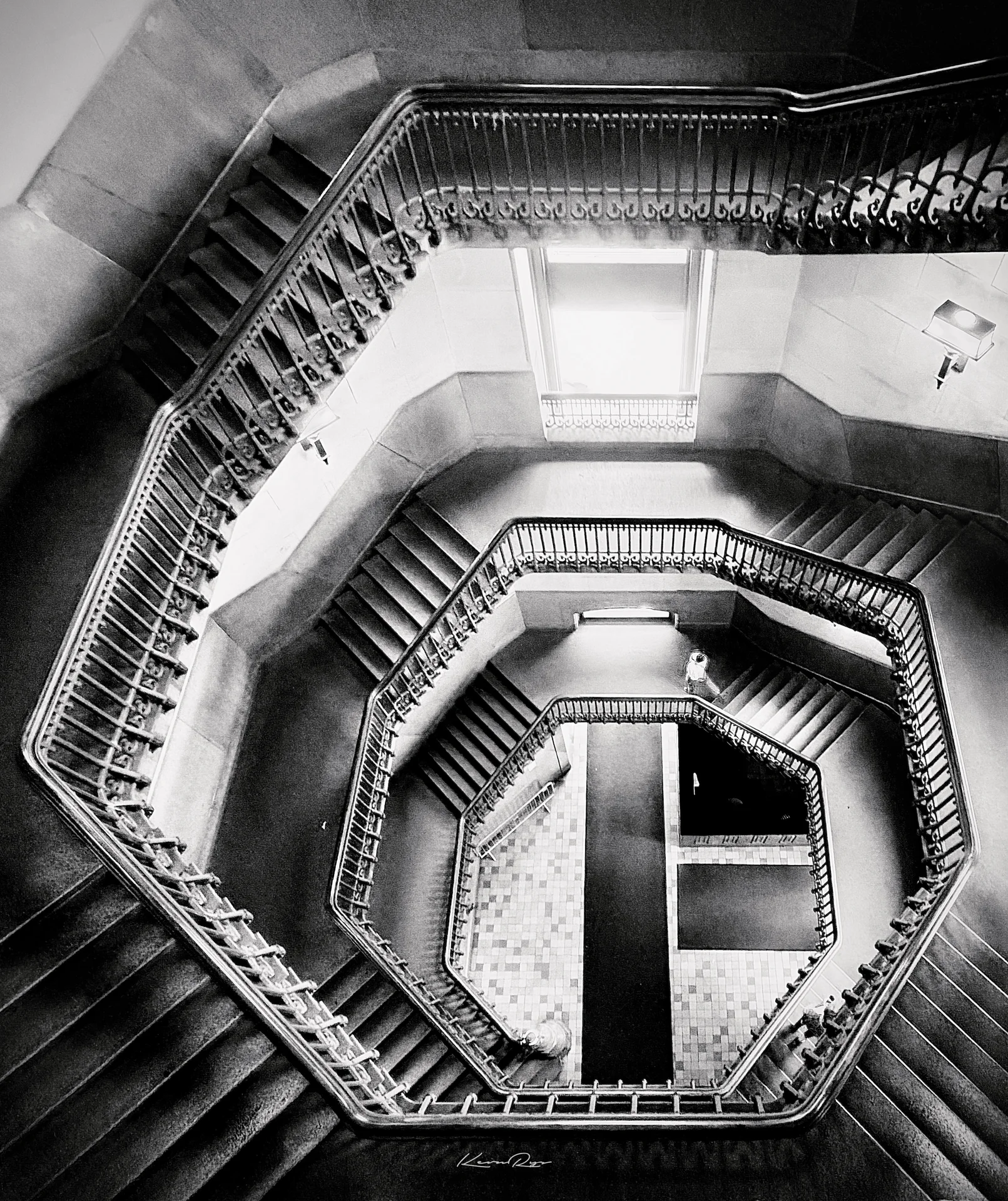 Looking straight down an octagonal stairwell in black and white, ornate iron railings winding to a tiled floor far below