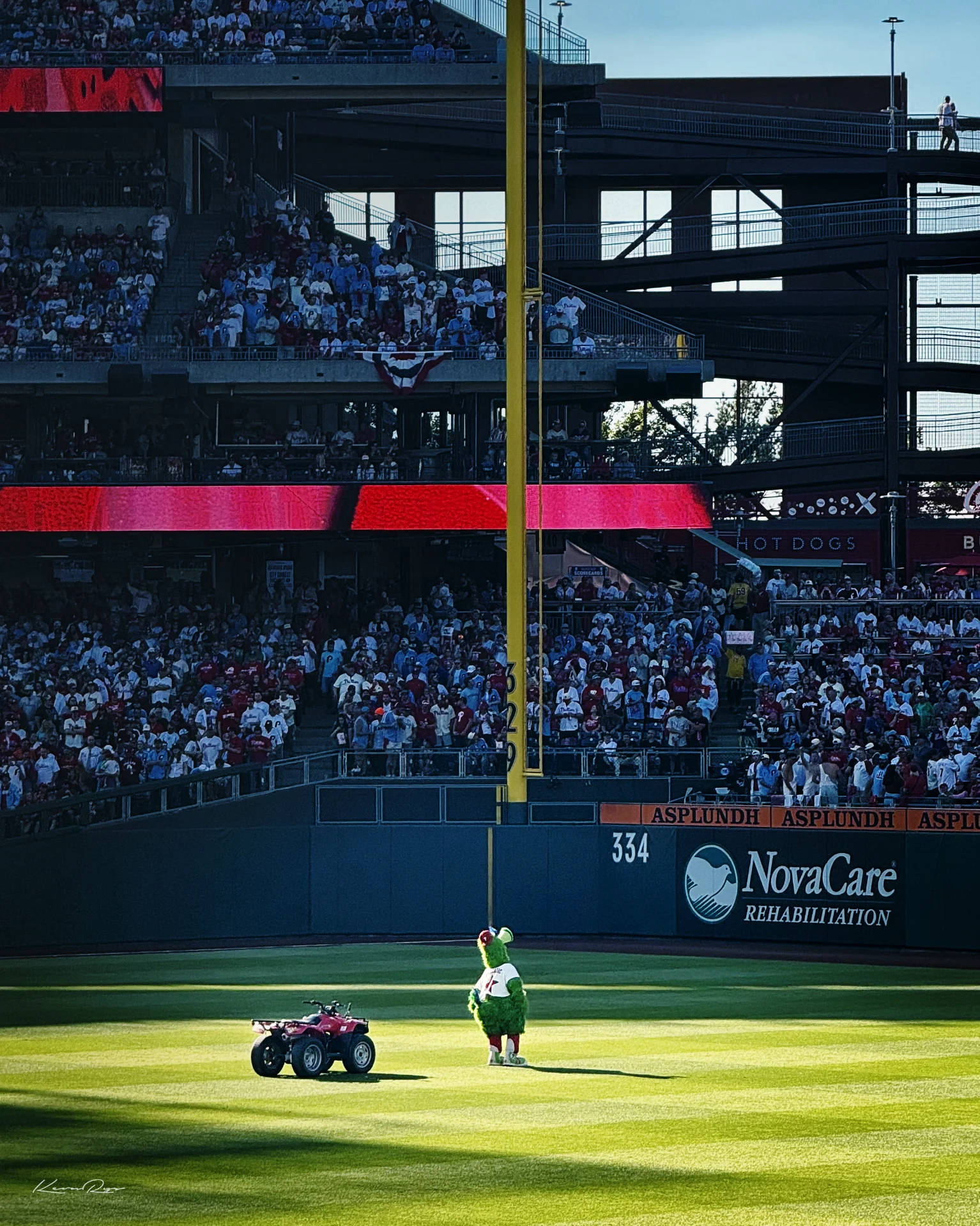 The Phanatic stands in the sunlit outfield next to a red four-wheeler, the yellow foul pole plus packed stands behind