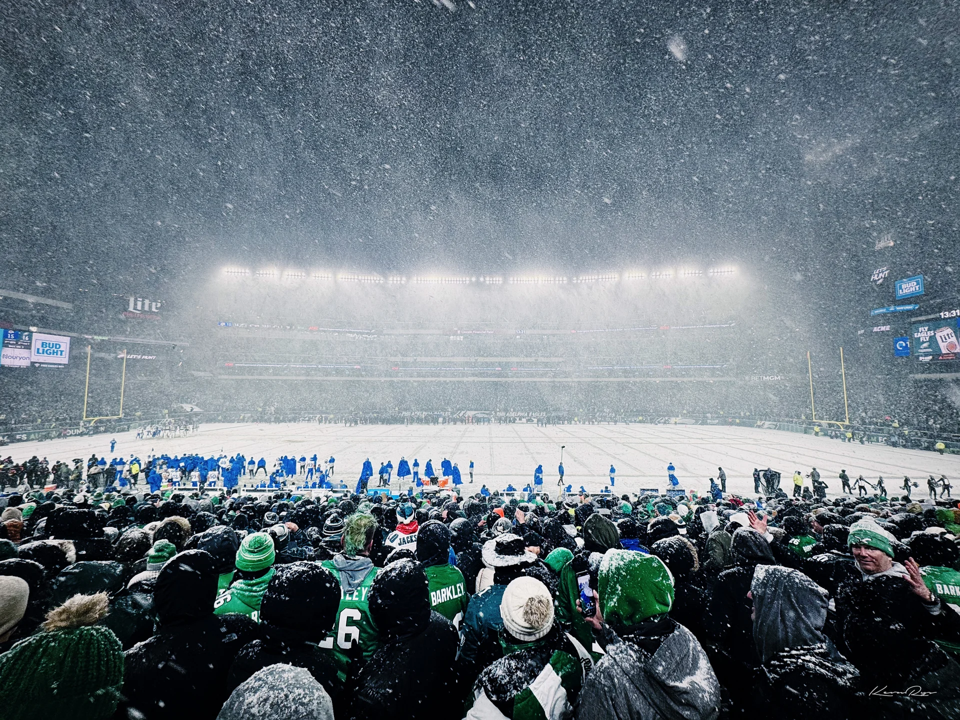 Heavy snow falls on a white field under stadium lights, players on the sideline, fans in green packed in the foreground