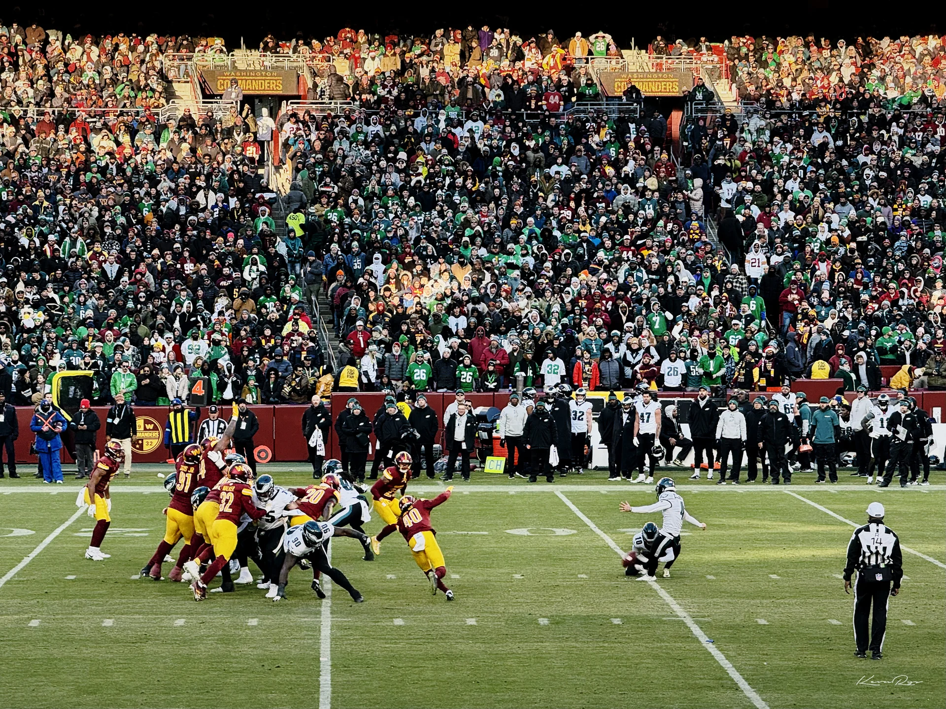 From the stands, a kicker swings at the held ball as players in burgundy and gold leap to block, a crowd full of green behind