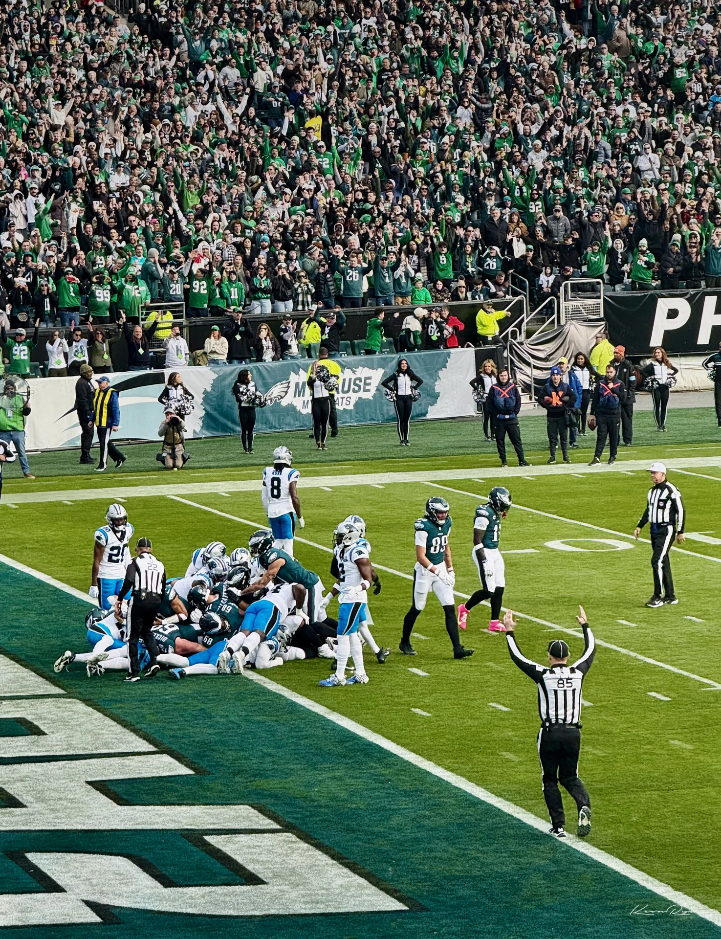 An official raises both arms as players pile up at the goal line, fans in green cheering in the stands behind
